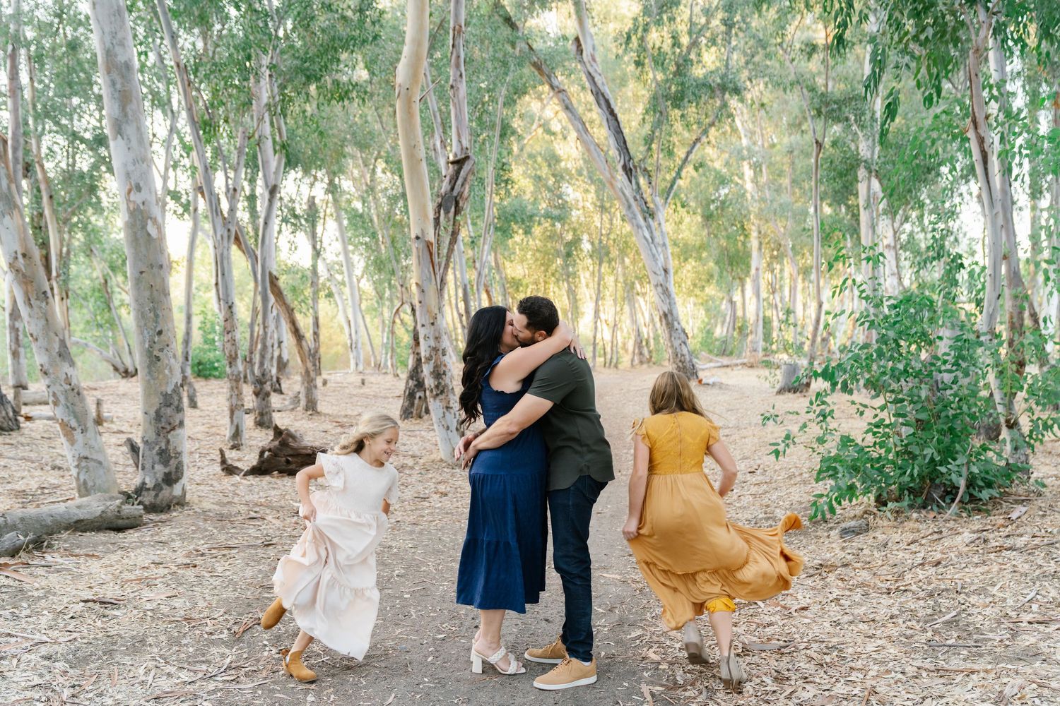 A family shares a tender kiss in a sunlit eucalyptus grove while two young daughters twirl around them.