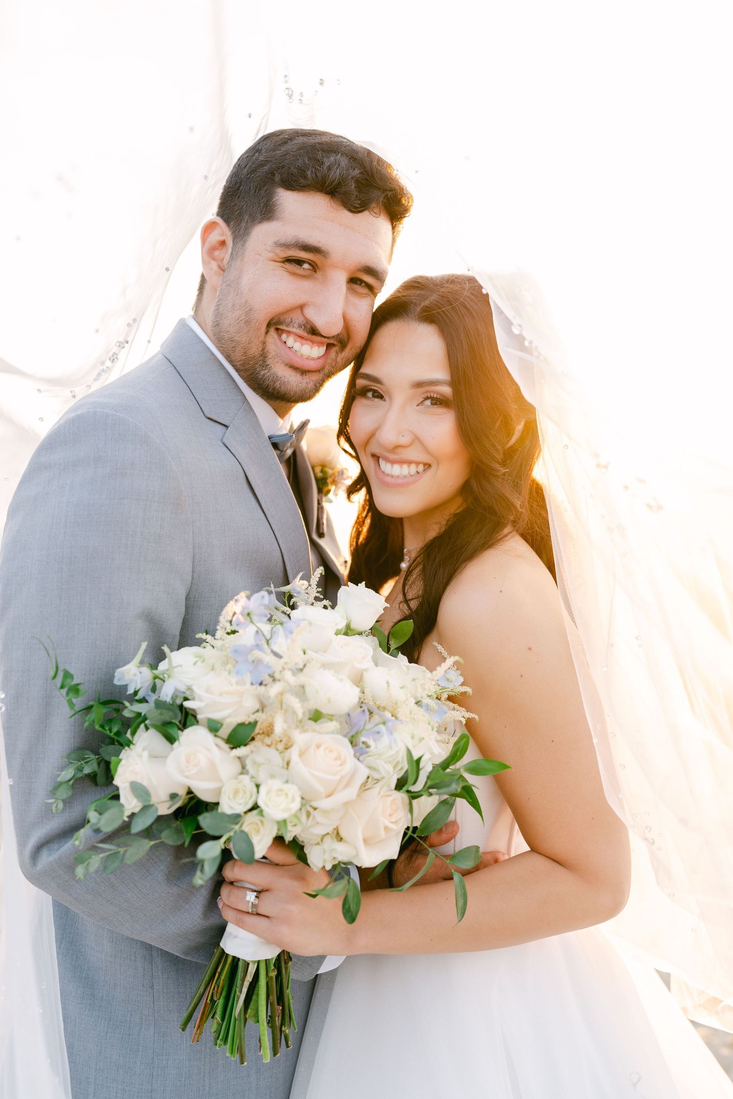 A wedding couple poses with a beautiful white floral bouquet against a light colored wall.