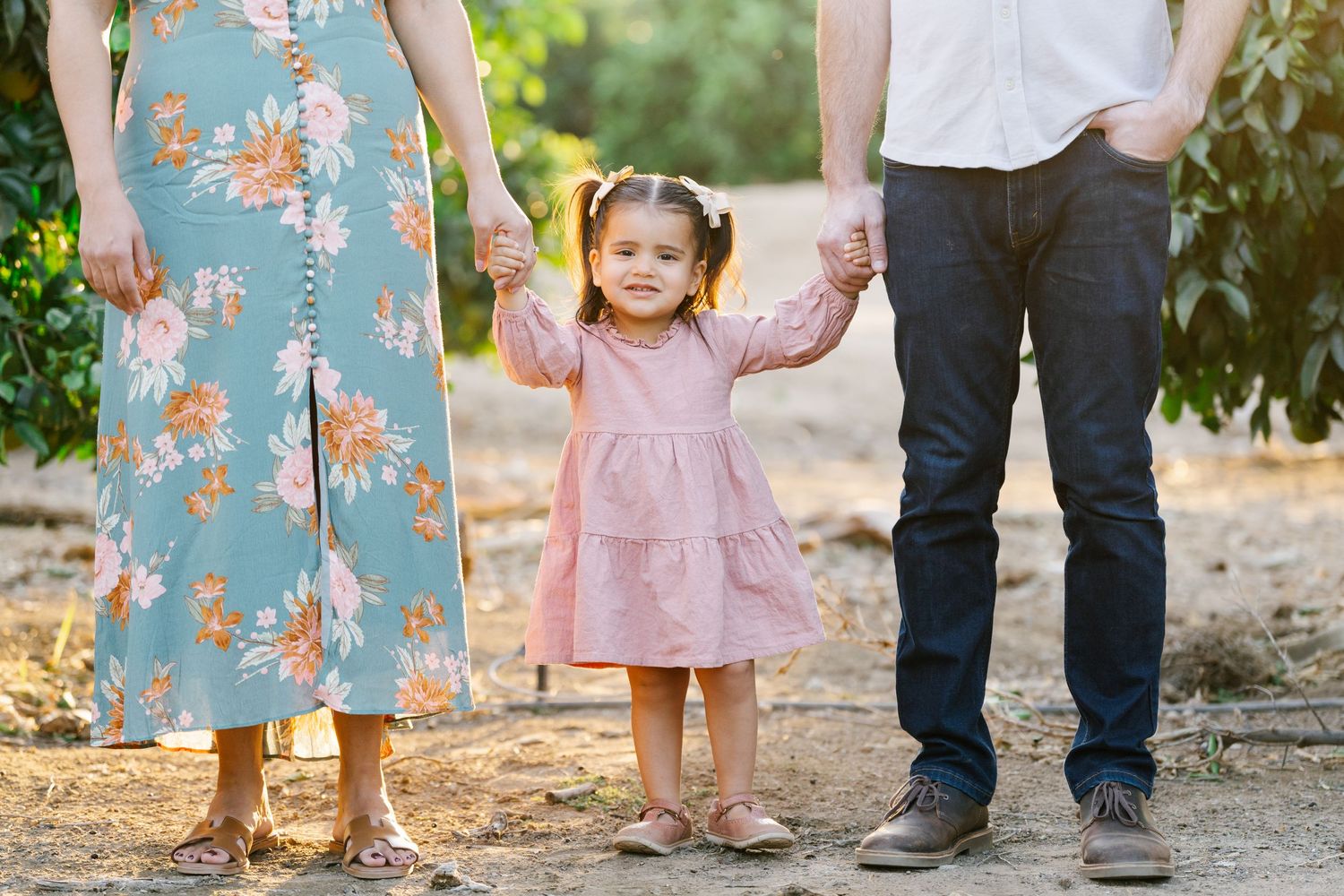 A family holds hands with their little one in a pink dress while walking through an orchard.