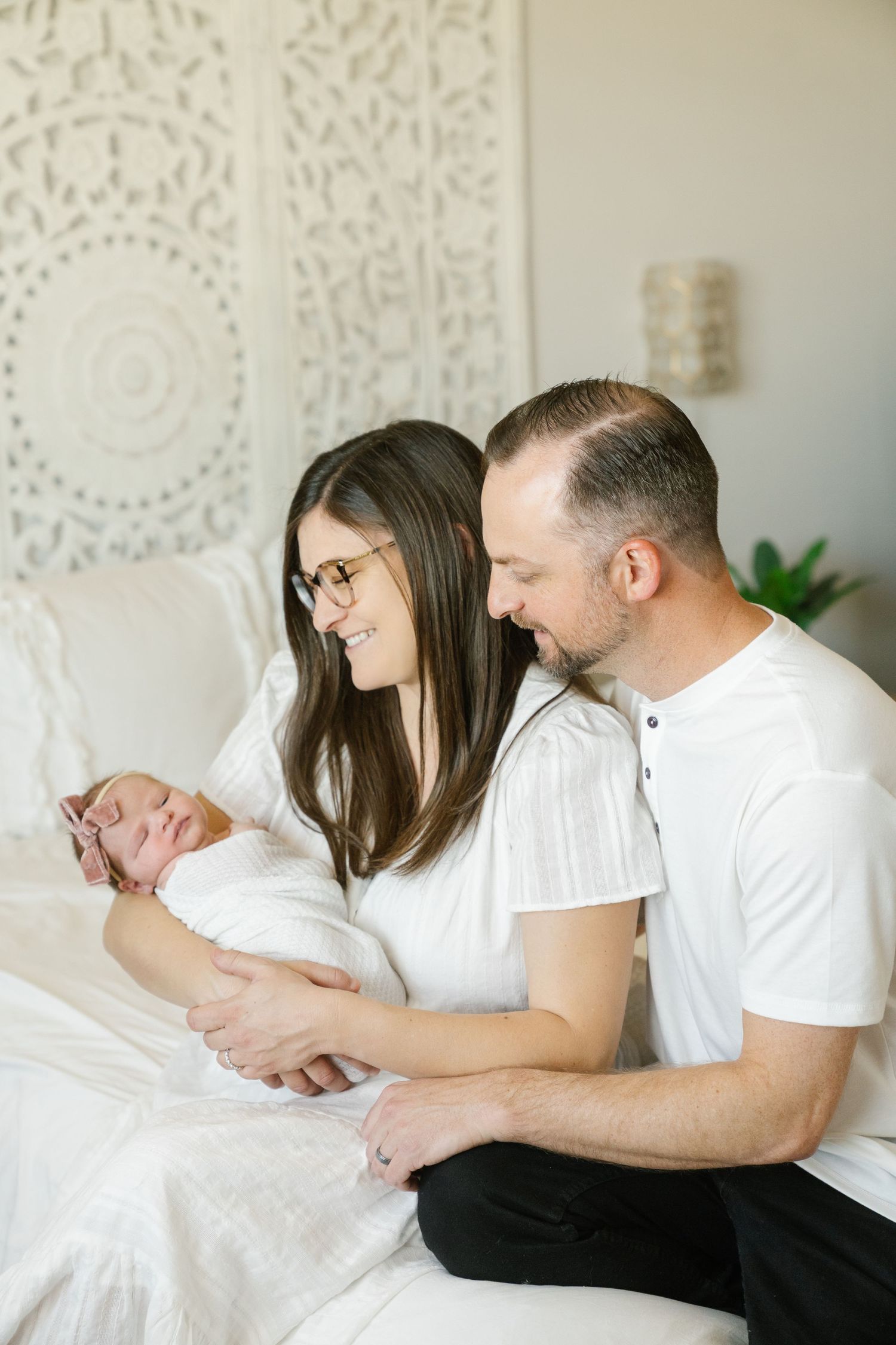 A tender moment as new parents cuddle their newborn baby together on a white bed with a decorative wall backdrop.