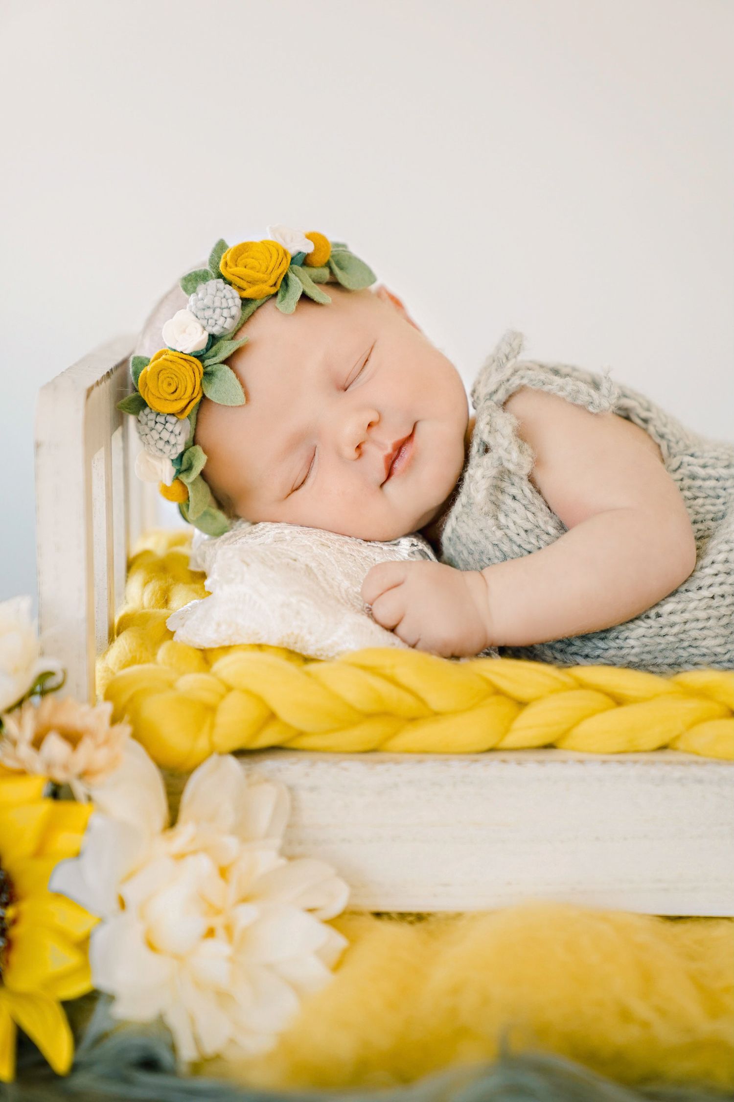 A sleeping baby wearing yellow floral crown rests on yellow bedding with flowers.