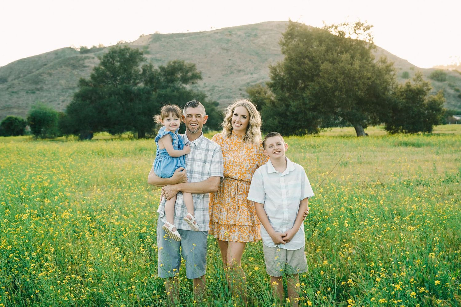 Family of four posing together in field of yellow wildflowers with mountains in background.