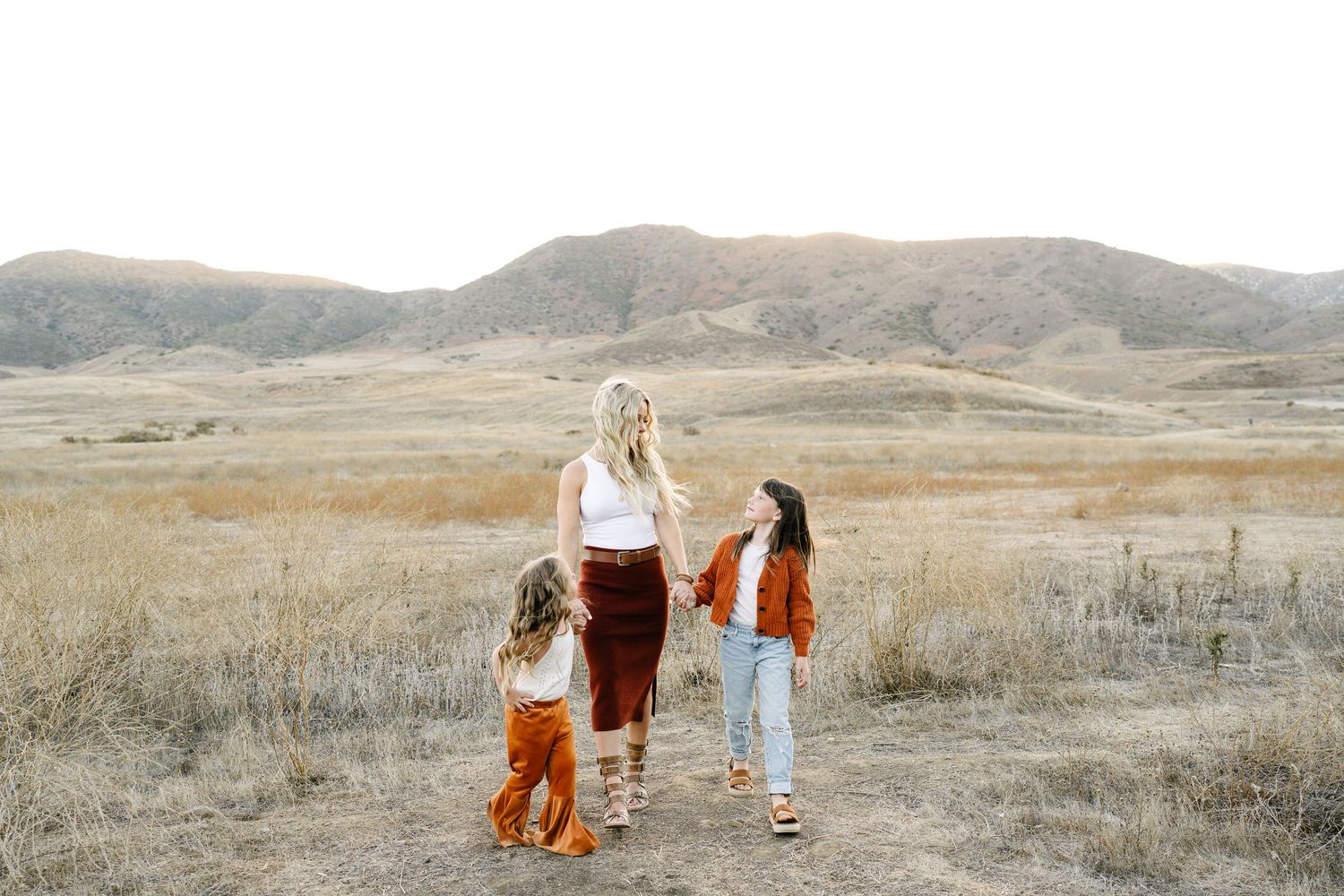 A mother and two children stand on a dirt path surrounded by golden fields and rolling hills.
