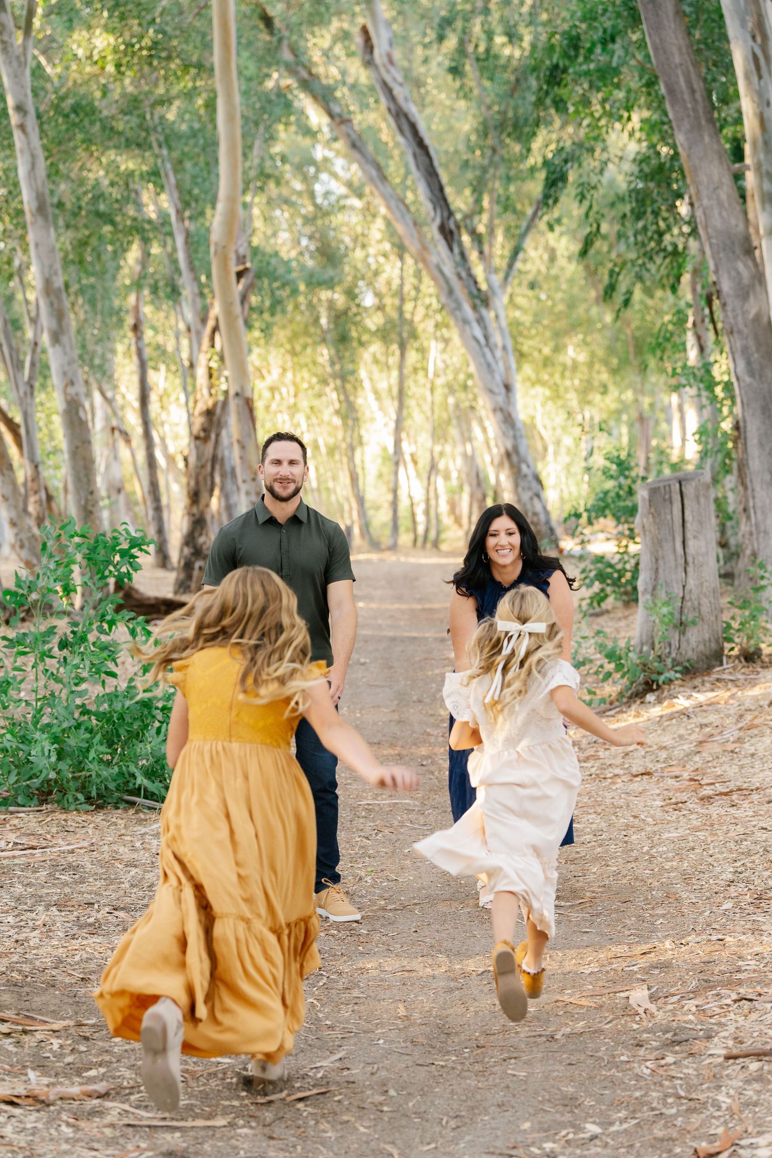 Family of four enjoying a sunny walk on a forest path, with two girls in dresses running toward their smiling parents.