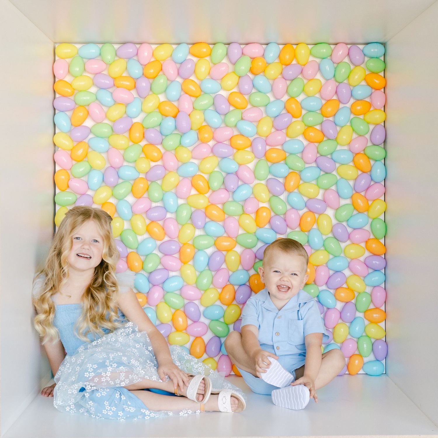 Two children sit against a colorful backdrop of pastel Easter eggs during a springtime photo session.