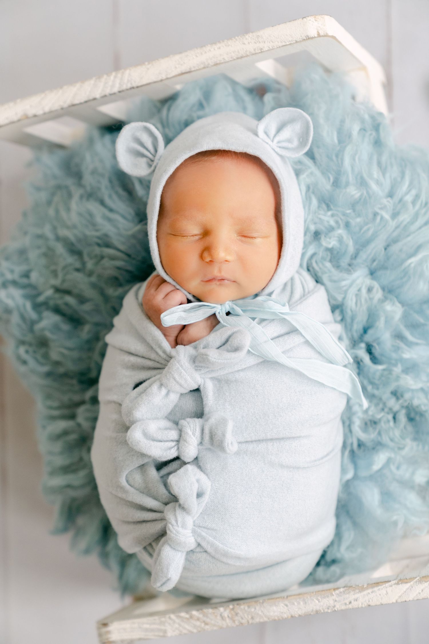 Sleeping newborn in white bear ear bonnet swaddled on blue fuzzy backdrop.
