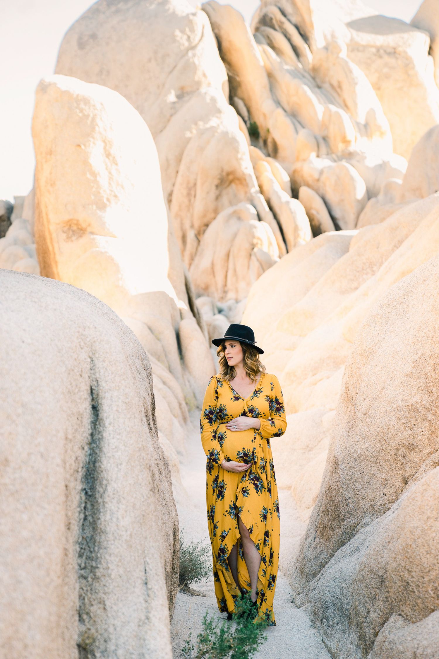 Woman in flowing yellow floral dress poses among dramatic white rock formations in desert setting.