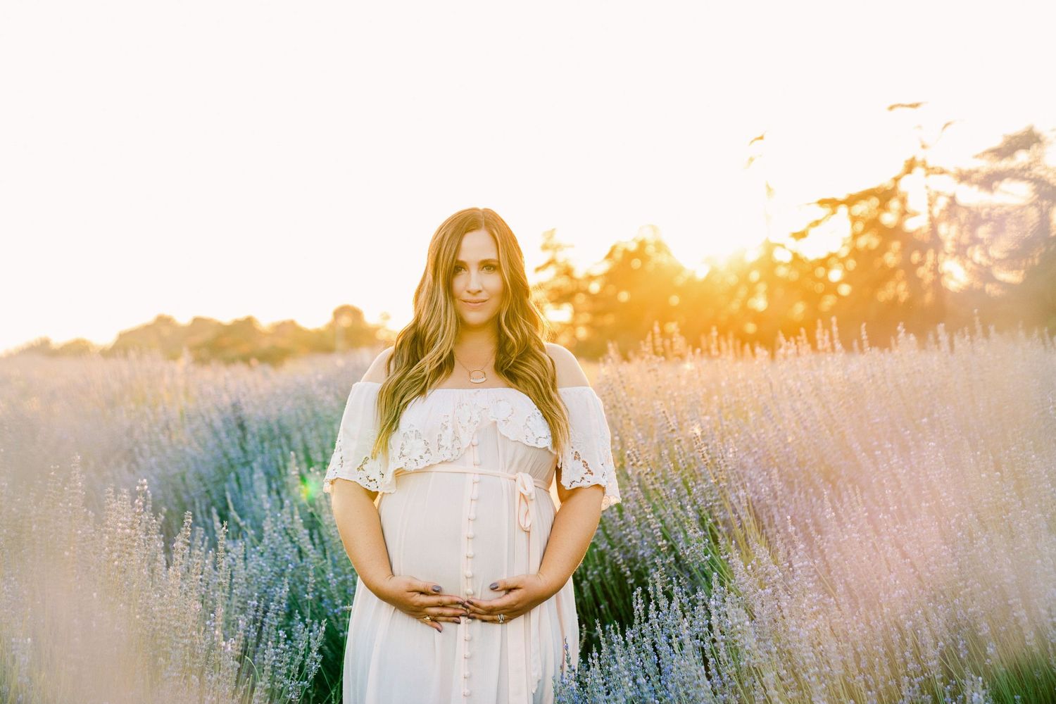 An expectant mother in white dress standing in a lavender field at sunset.