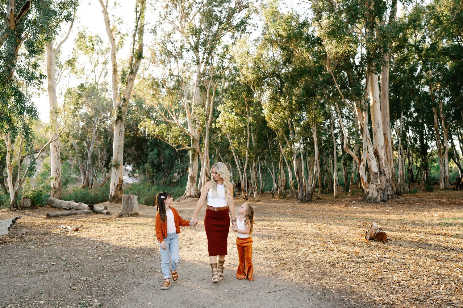 A woman walks hand-in-hand with two children through a sunlit eucalyptus grove on a dirt path.