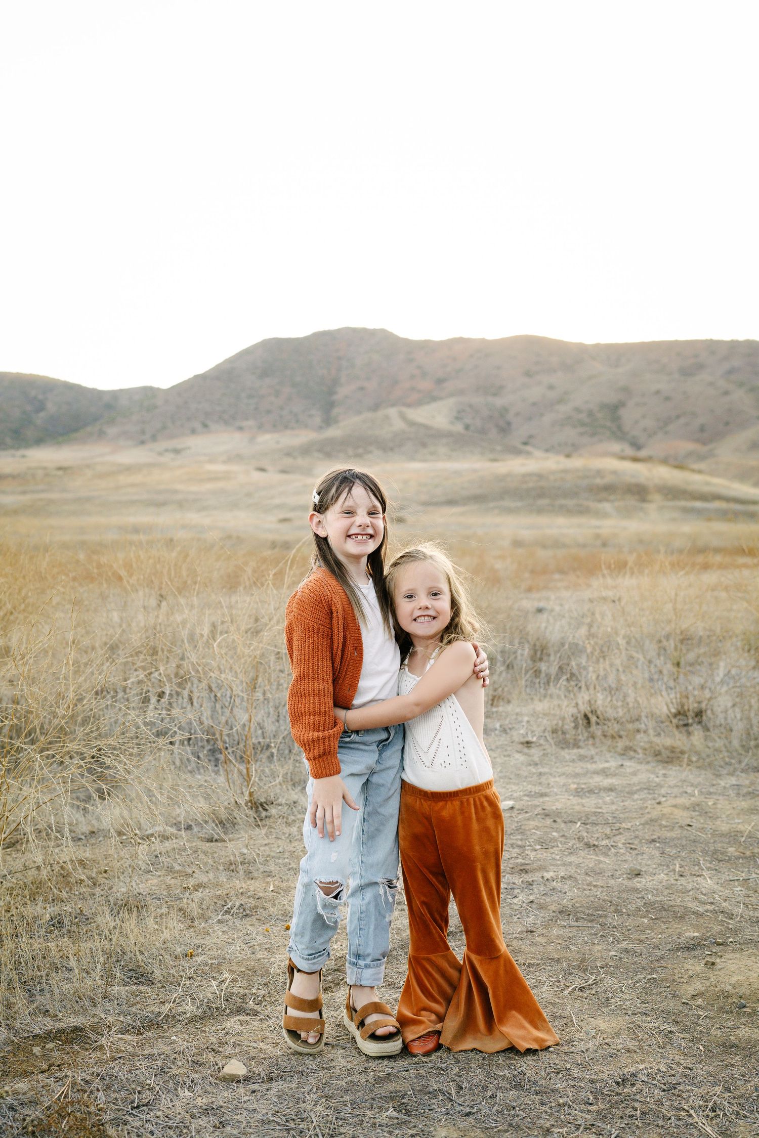 Two young girls hugging and smiling in a golden field with rolling hills backdrop.