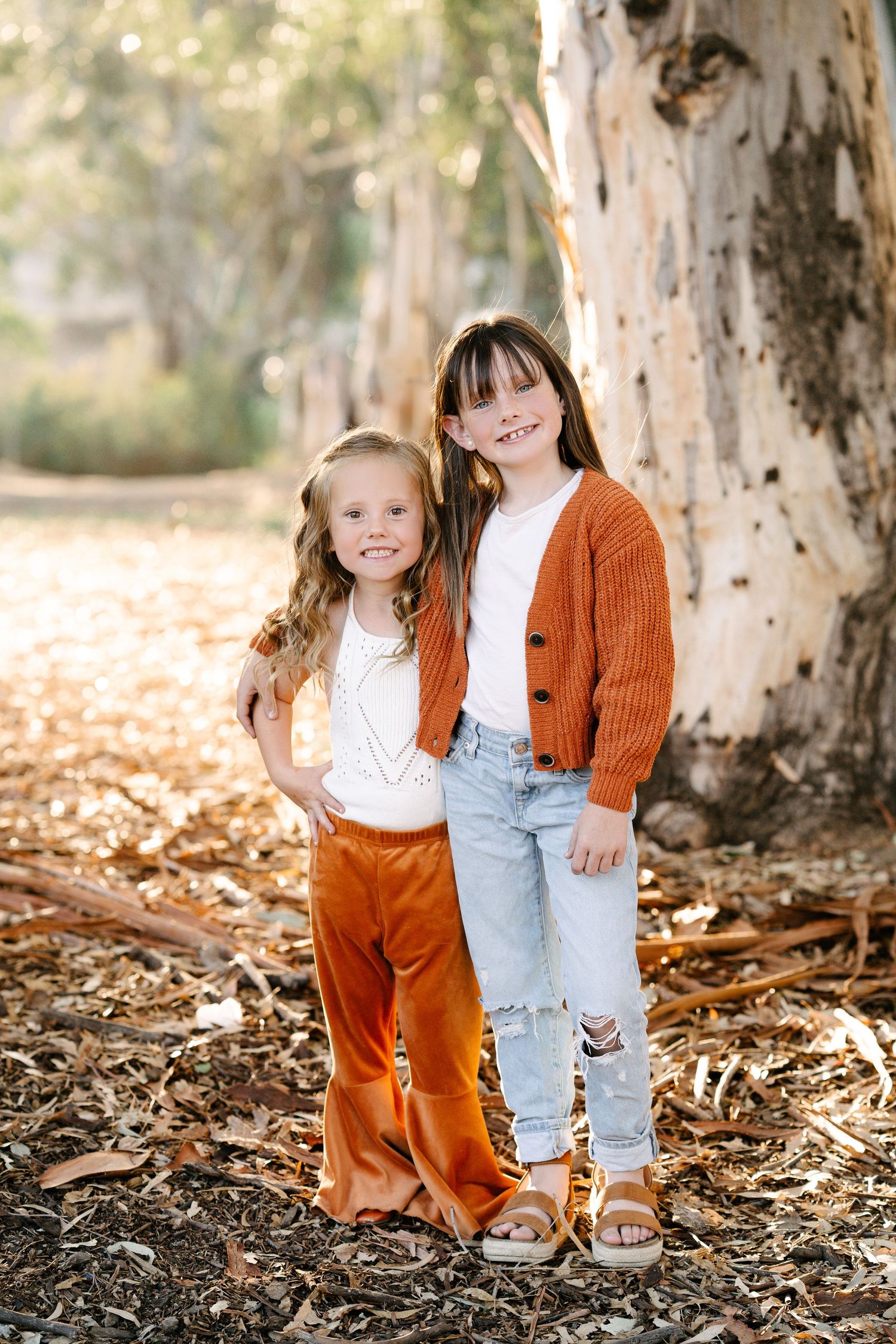 Two young girls in rust and orange outfits smile together beside a large tree trunk outdoors.
