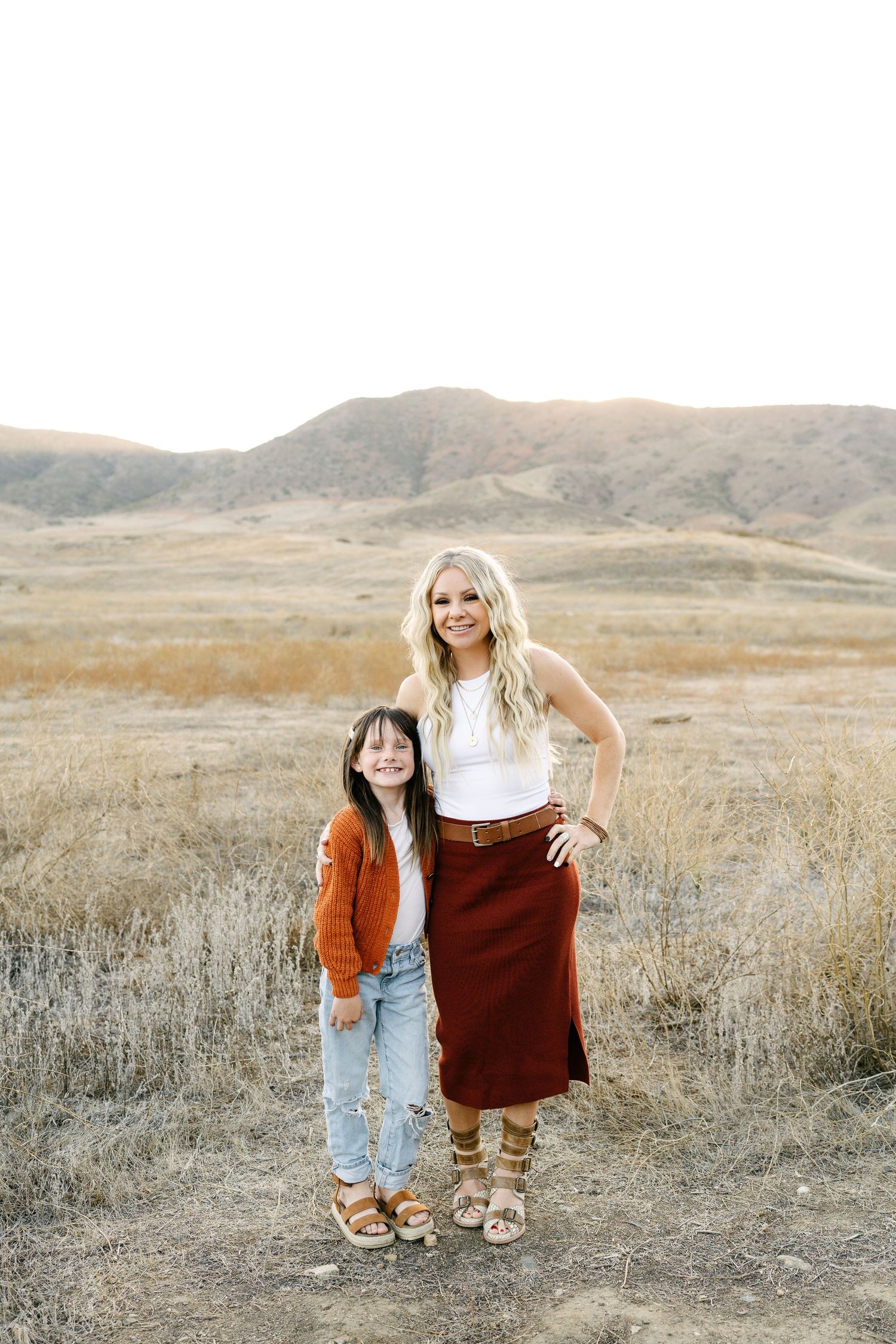 A blonde woman and young girl pose together in a golden field with hills behind.