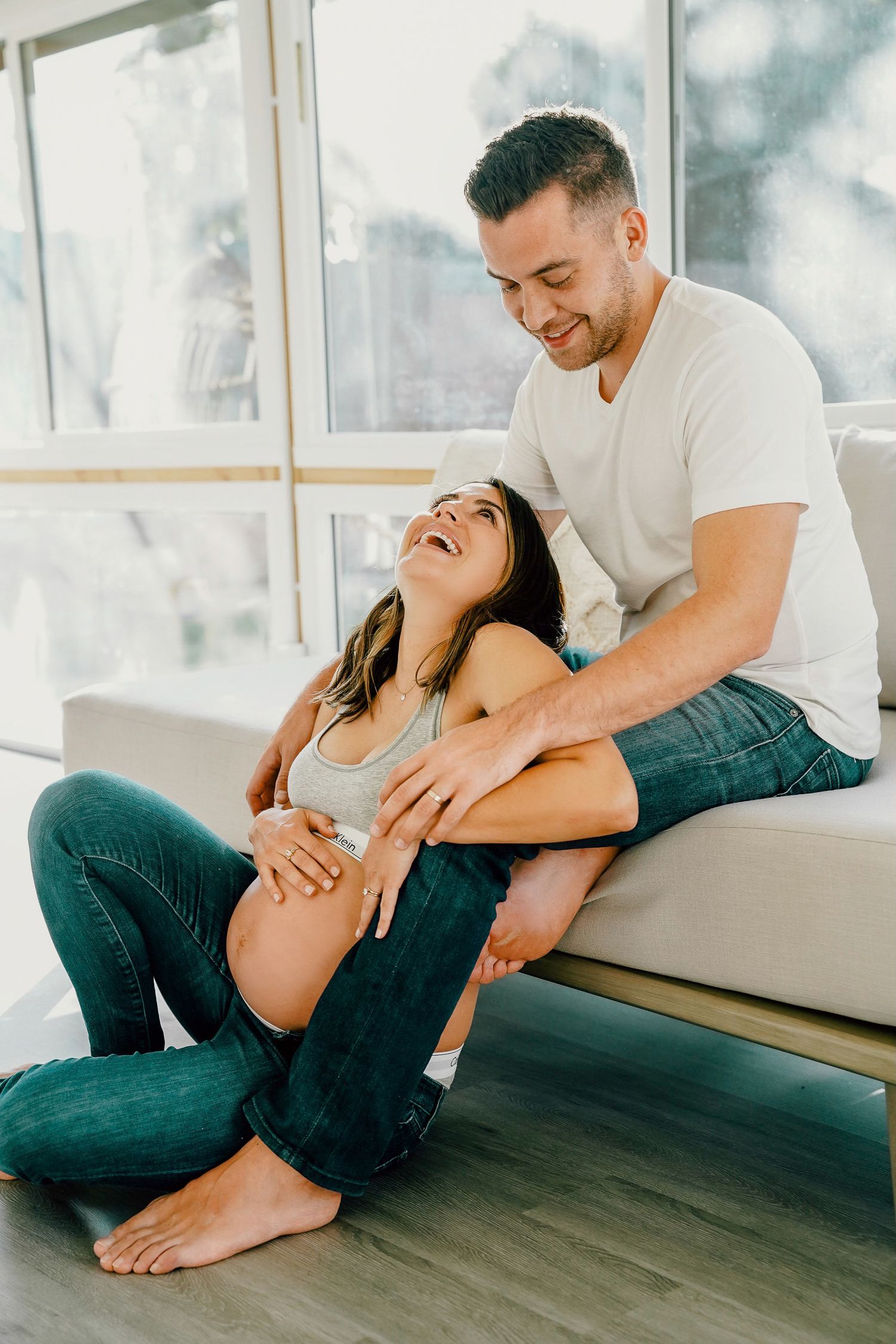 Intimate maternity photo of expecting couple sharing joyful moment together on couch in bright sunlit room.