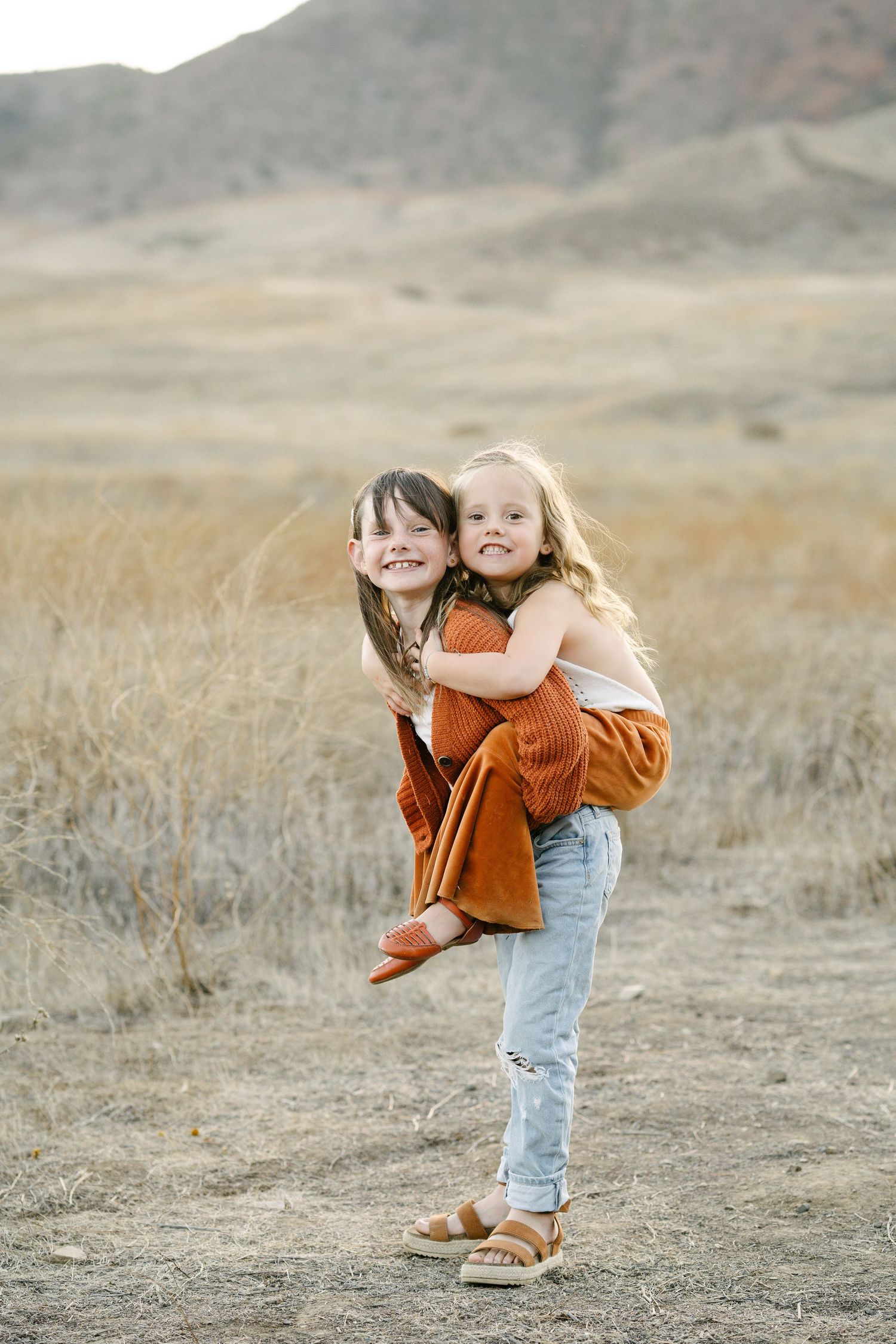 Two young sisters play piggyback in a dry golden field with rolling hills in the background.
