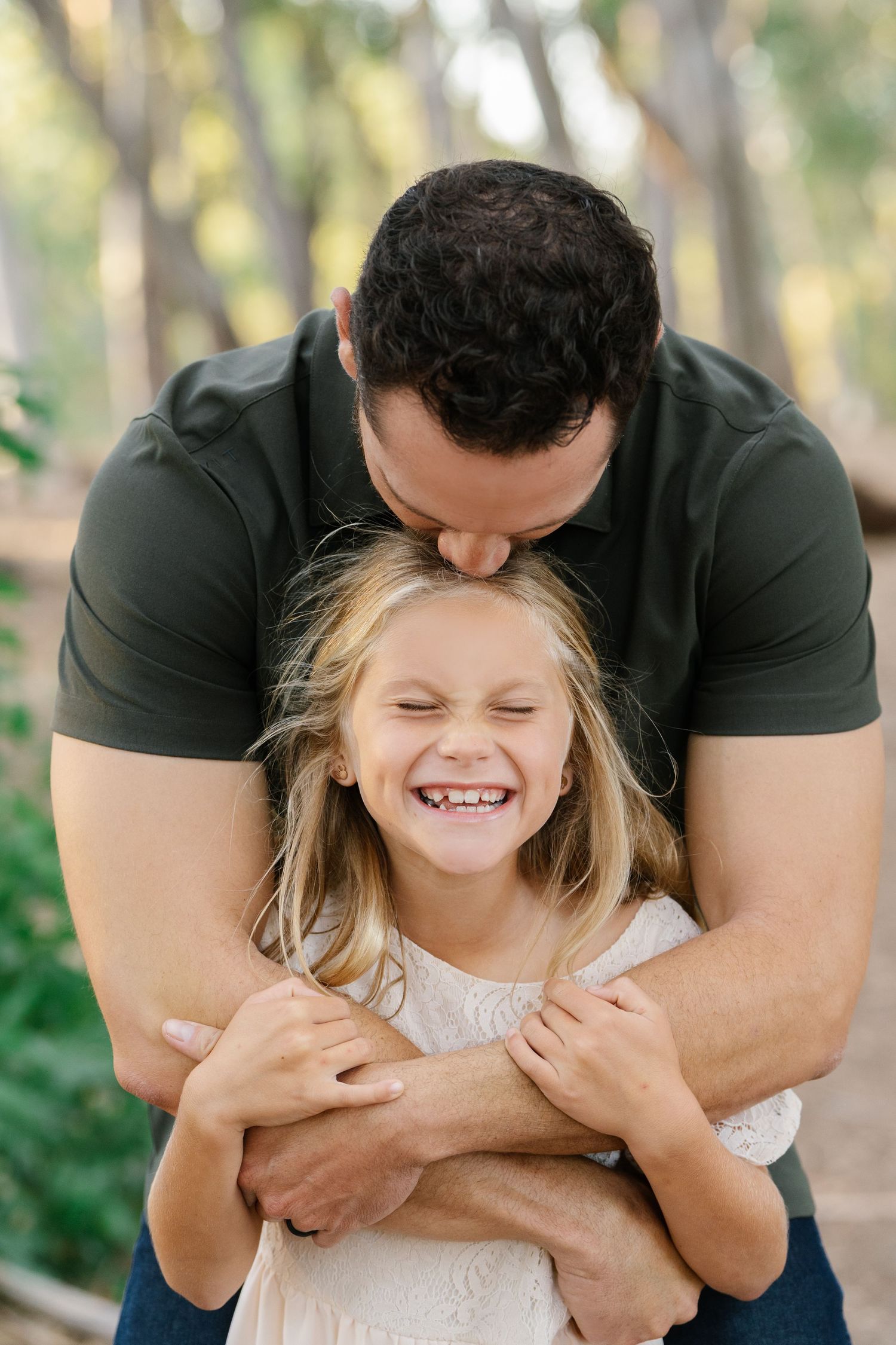 A father kisses his laughing young blonde girl on the head while hugging her outdoors among green trees.