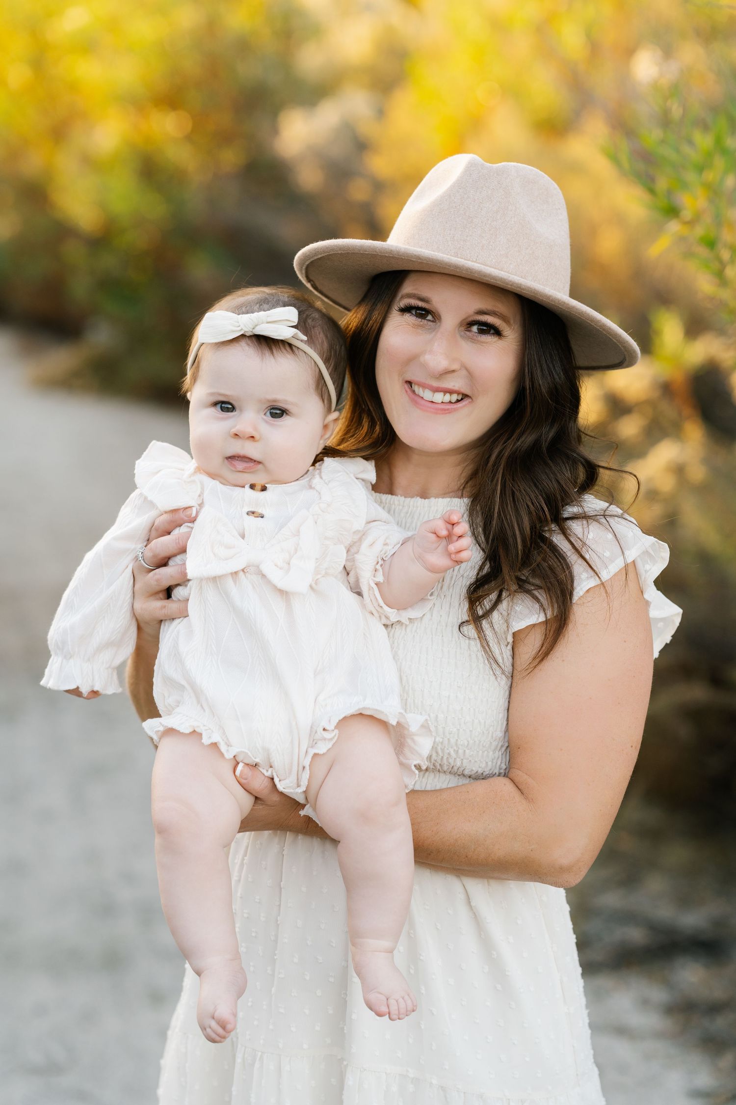 Woman in white dress and hat holding baby in matching white outfit against fall backdrop.