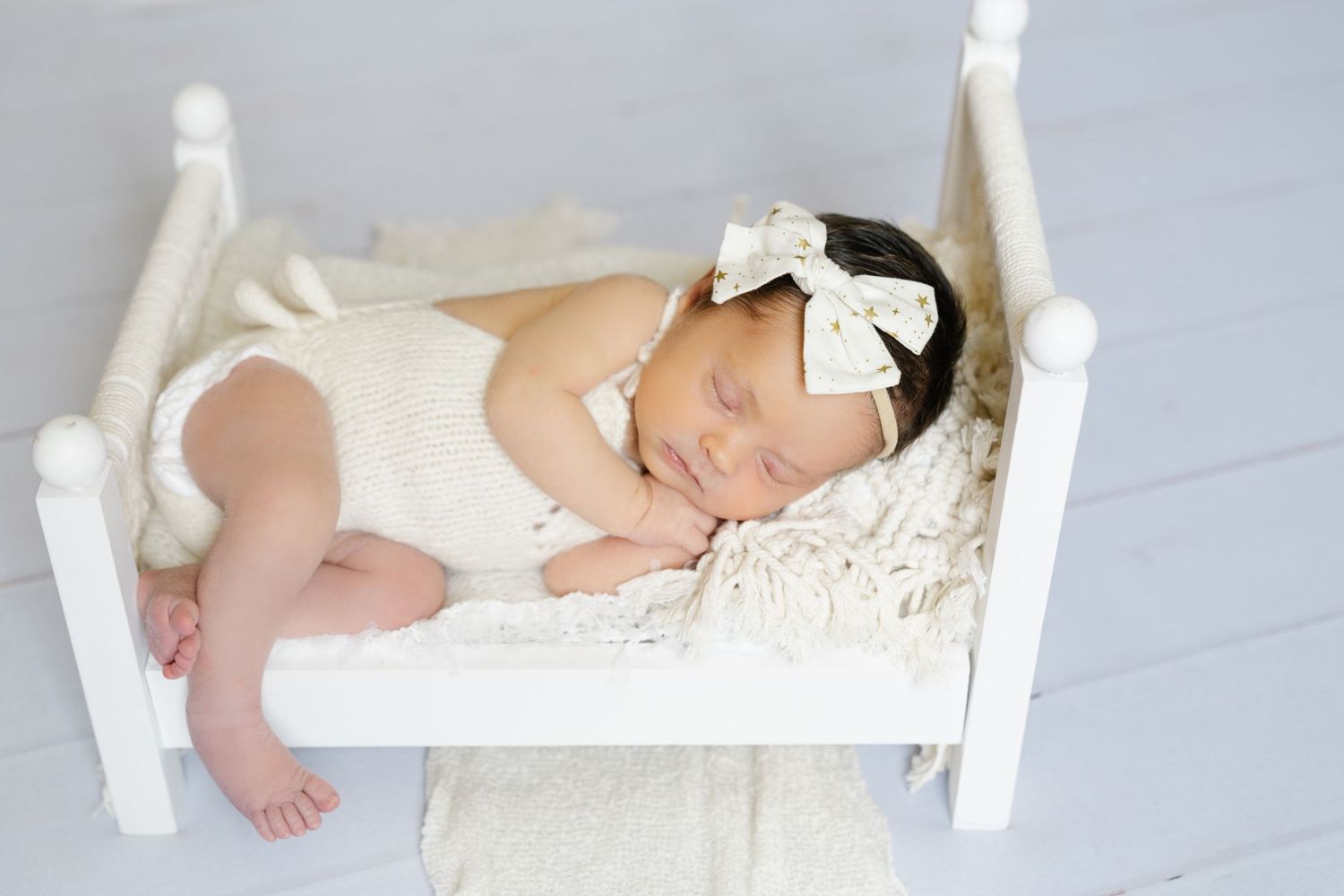 Sleeping newborn in white outfit laying on small white wooden bed with decorative bow.