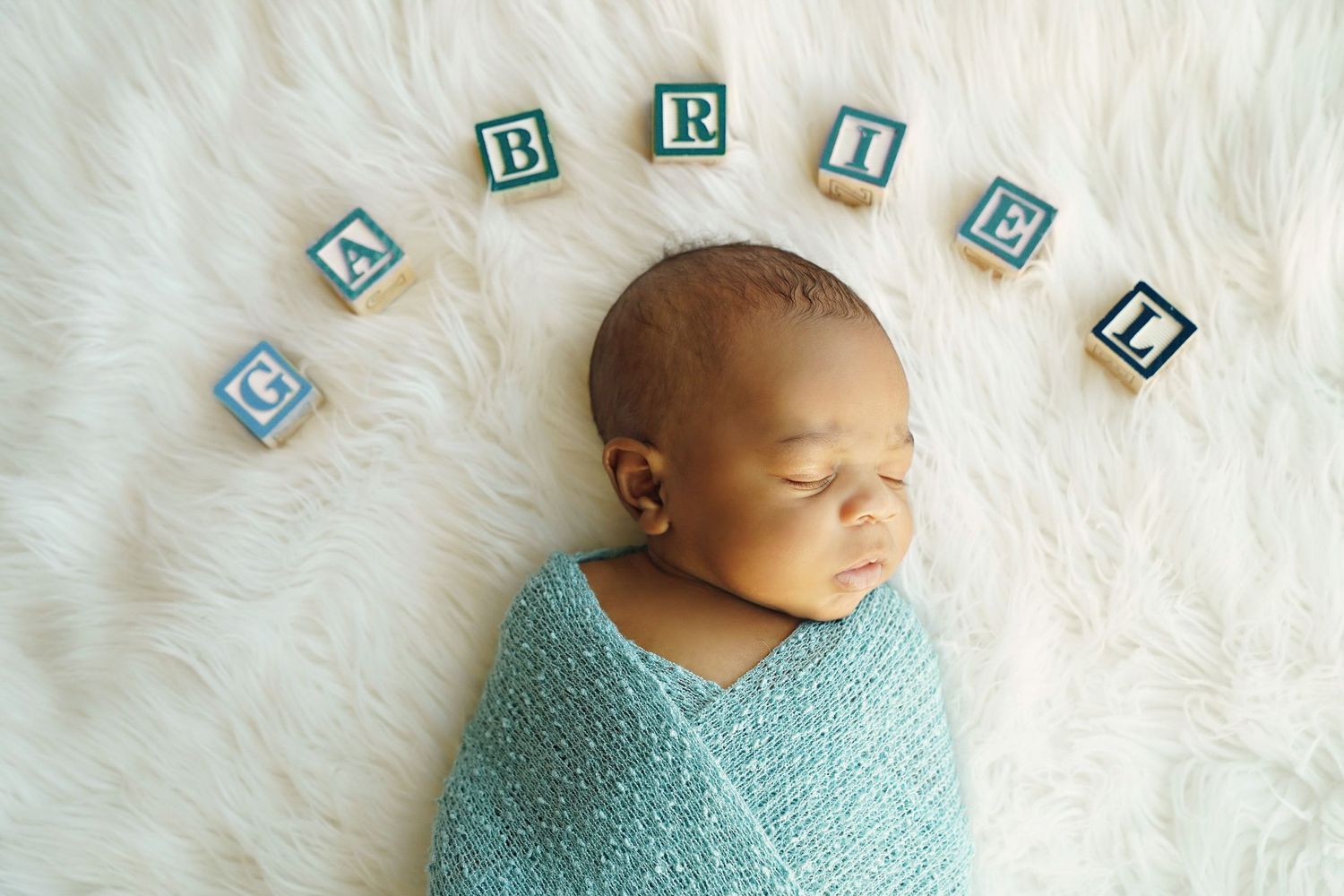 Newborn wrapped in blue swaddle blanket with wooden alphabet blocks spelling above.
