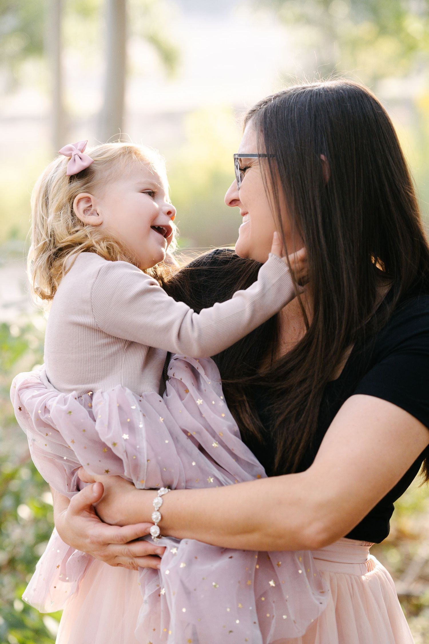 A smiling mother in black top holds her laughing toddler daughter in a pink tulle dress outdoors.