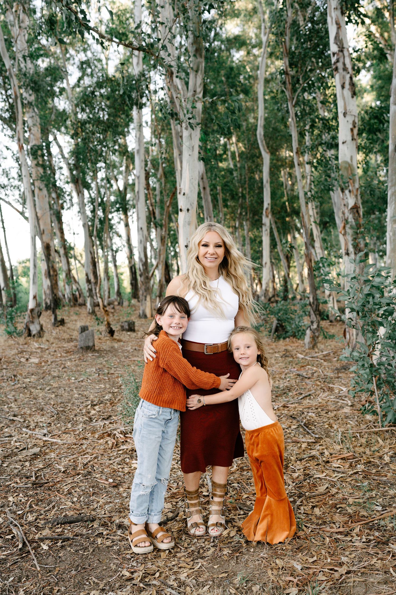 Pregnant woman in white top and rust skirt hugs two children in a sunlit eucalyptus forest setting.