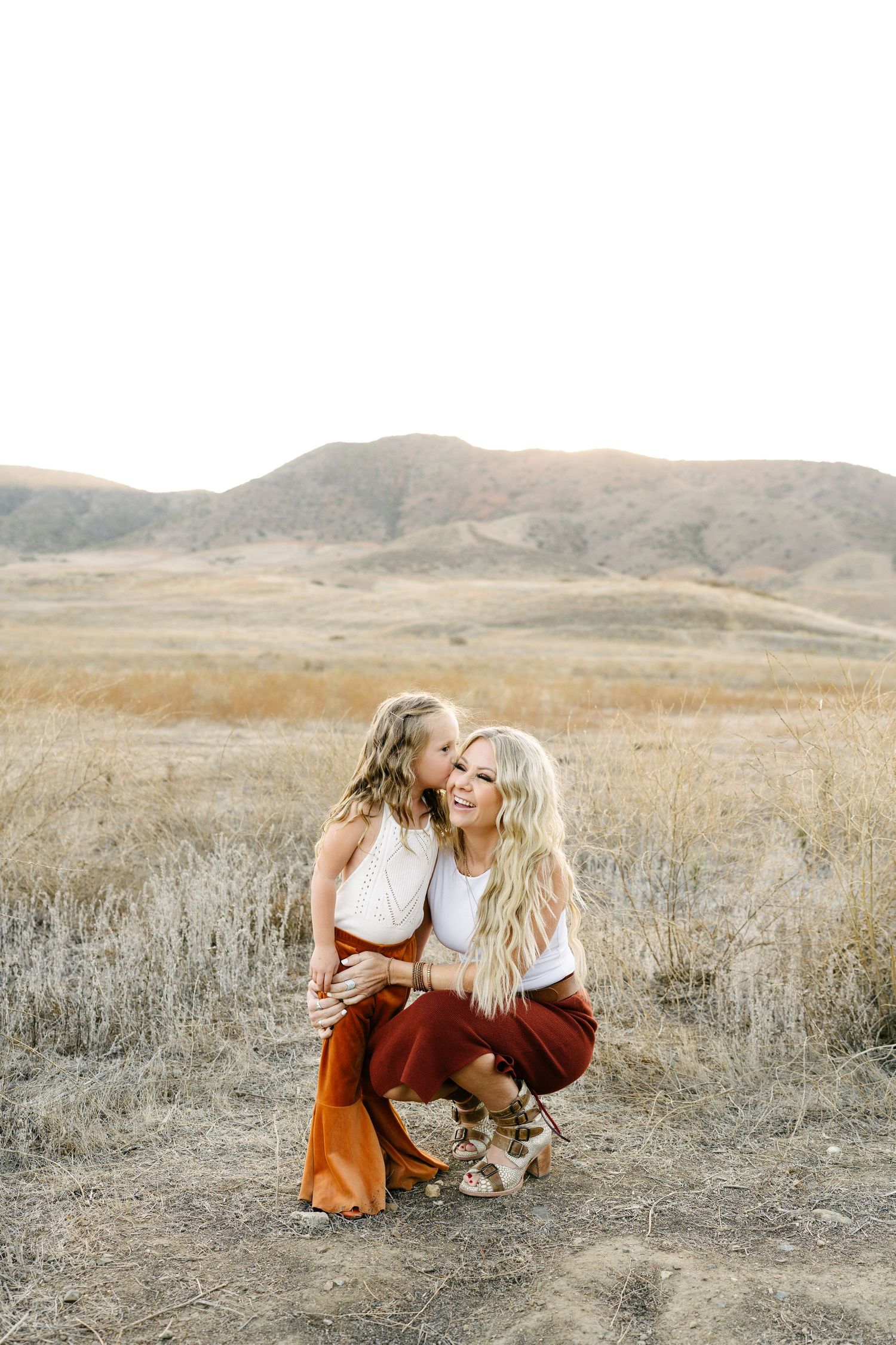 A mother crouches in a golden field as her young daughter gives her a sweet kiss on the cheek.