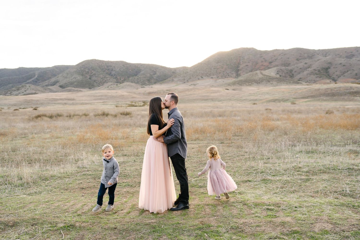 Family of four poses in golden field with rolling hills, parents sharing a kiss while two young children stand nearby.