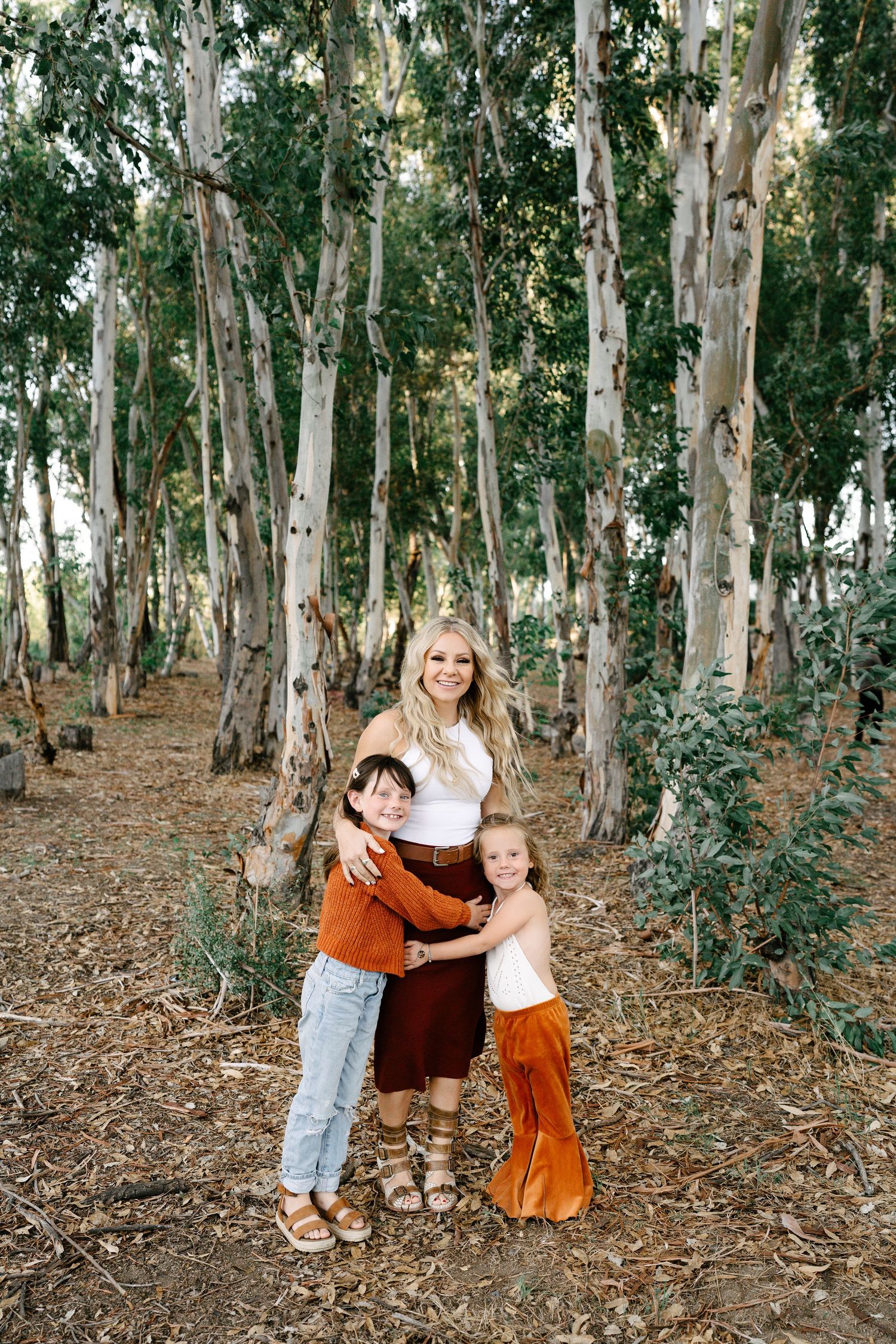 A blonde mother hugs her two children in an eucalyptus forest, wearing earth tones.