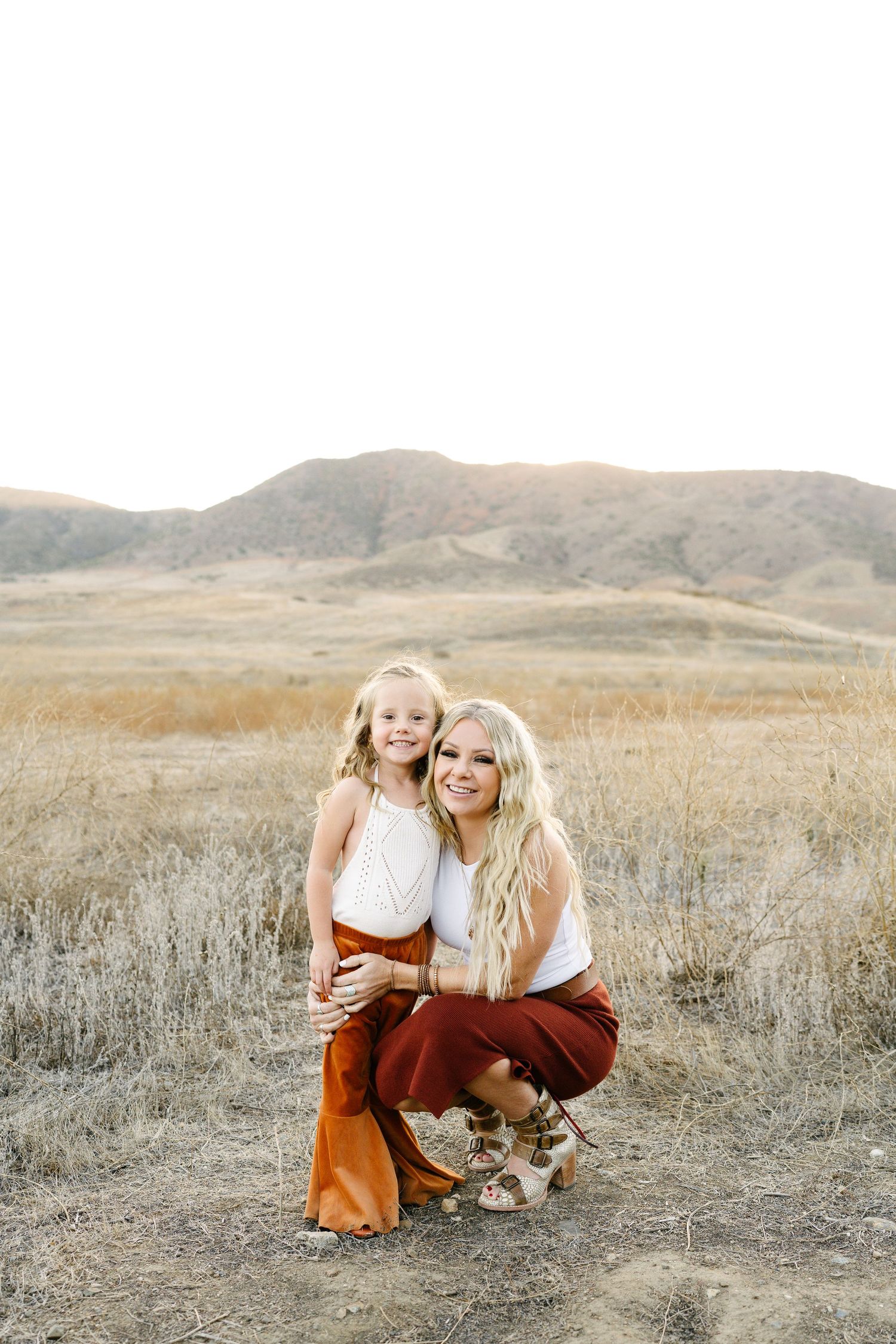 A blonde woman crouches beside a young girl in rust pants against a golden hillside backdrop.