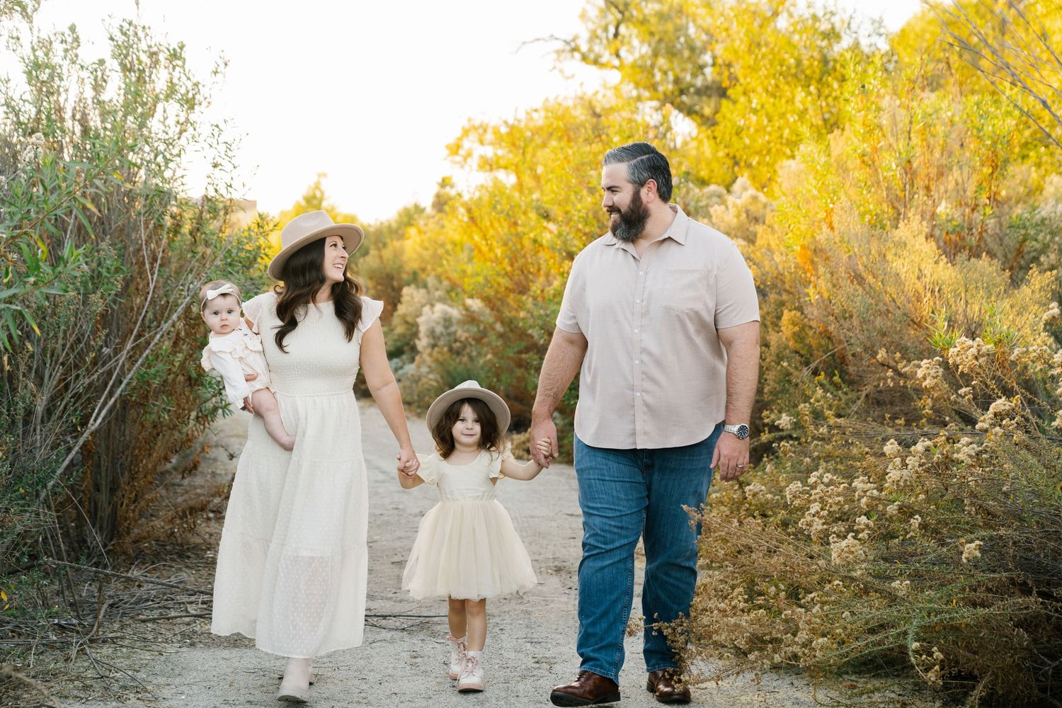 Family of four walking together on desert trail with golden sunlight filtering through bushes.
