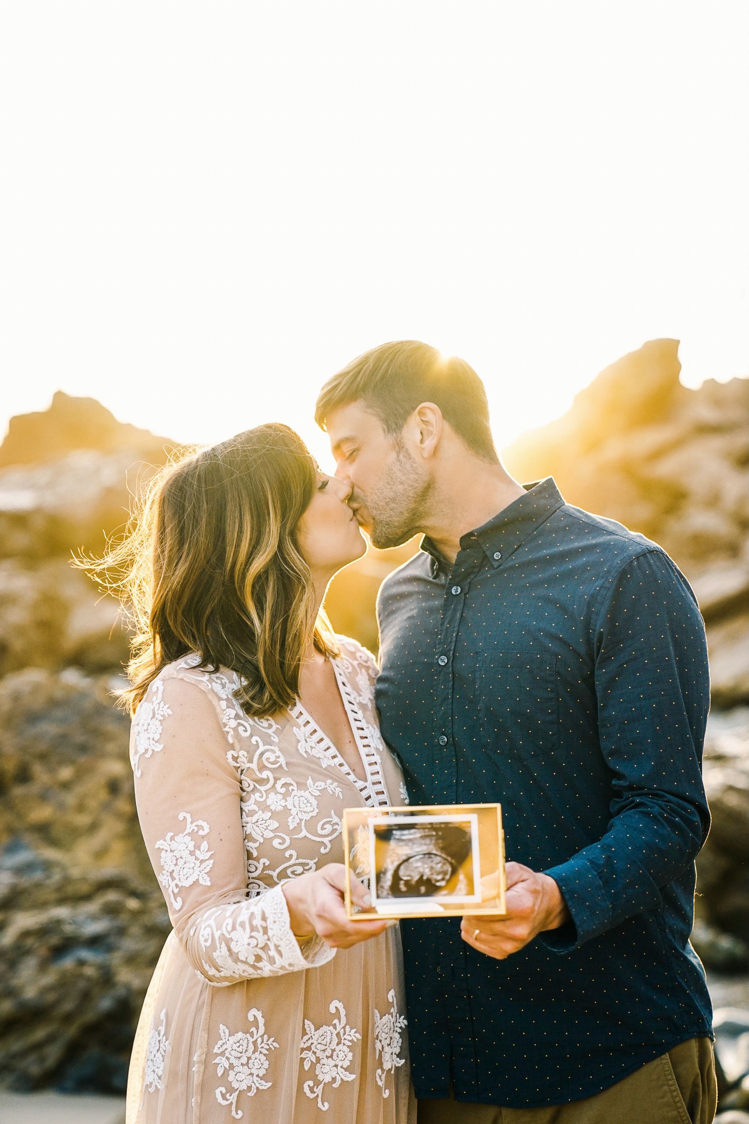 Sunset maternity photoshoot with couple holding ultrasound photo on rocky beach.