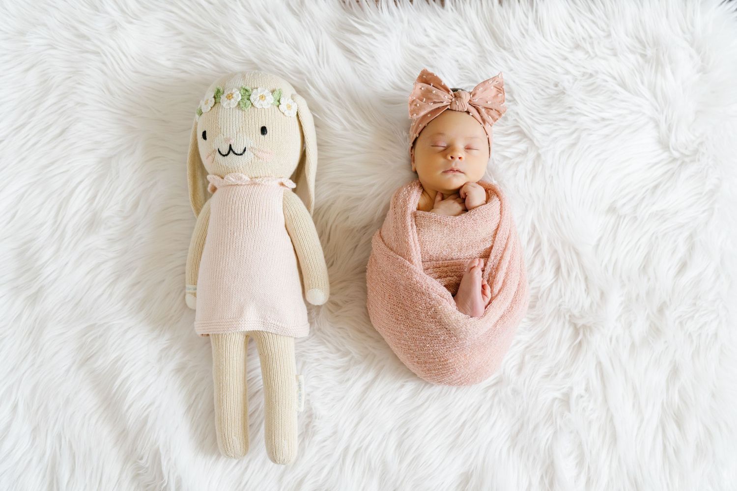Newborn baby in pink wrap next to matching stuffed bunny doll on white fur background.