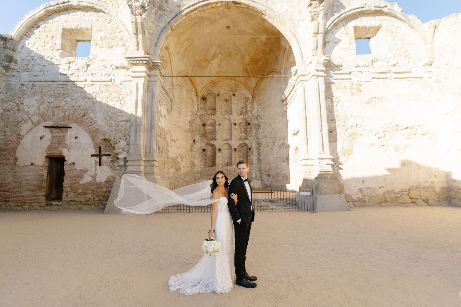 Newlyweds posing at ancient stone ruins with flowing white wedding veil.