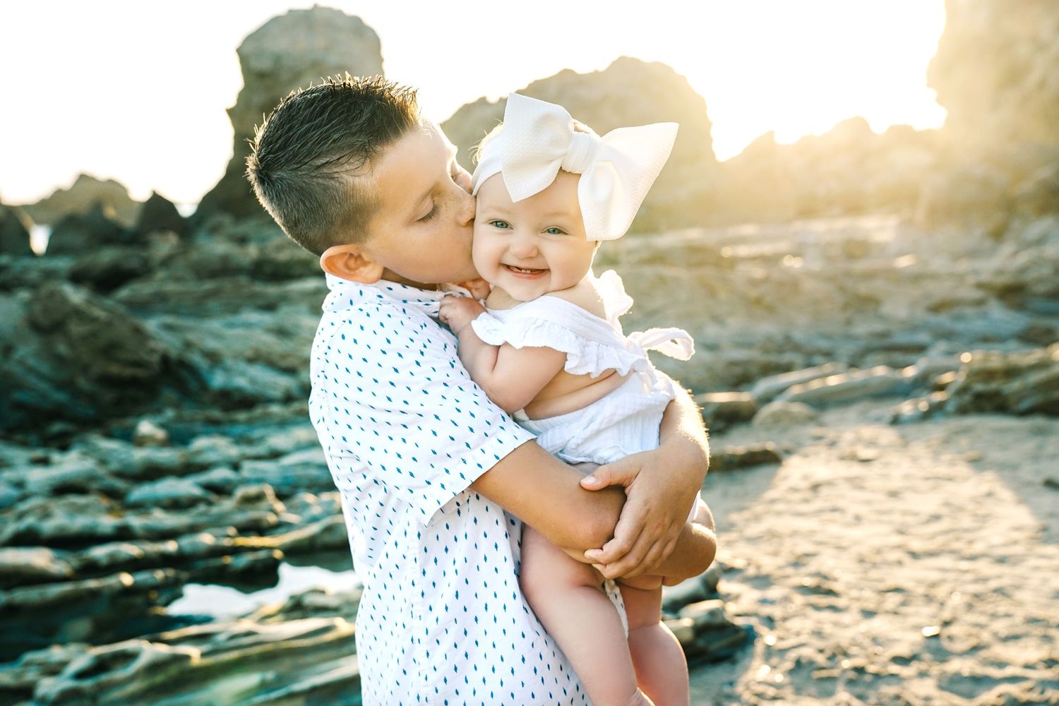 A tender moment shared between parent and baby against a rocky beach backdrop during golden hour.