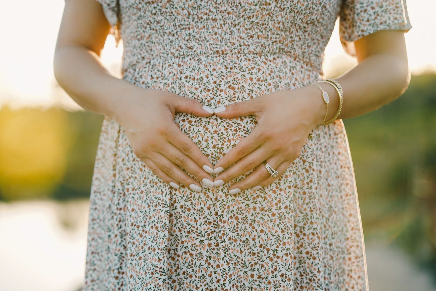 Close-up of hands forming heart shape over pregnant belly in floral dress during sunset.