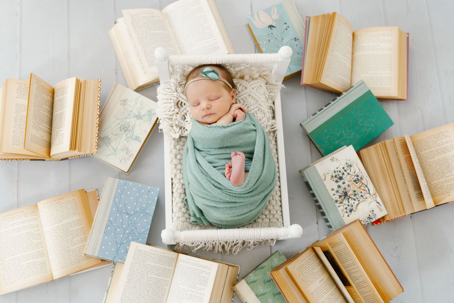 Sleeping newborn wrapped in teal swaddle surrounded by open books in soft lighting.