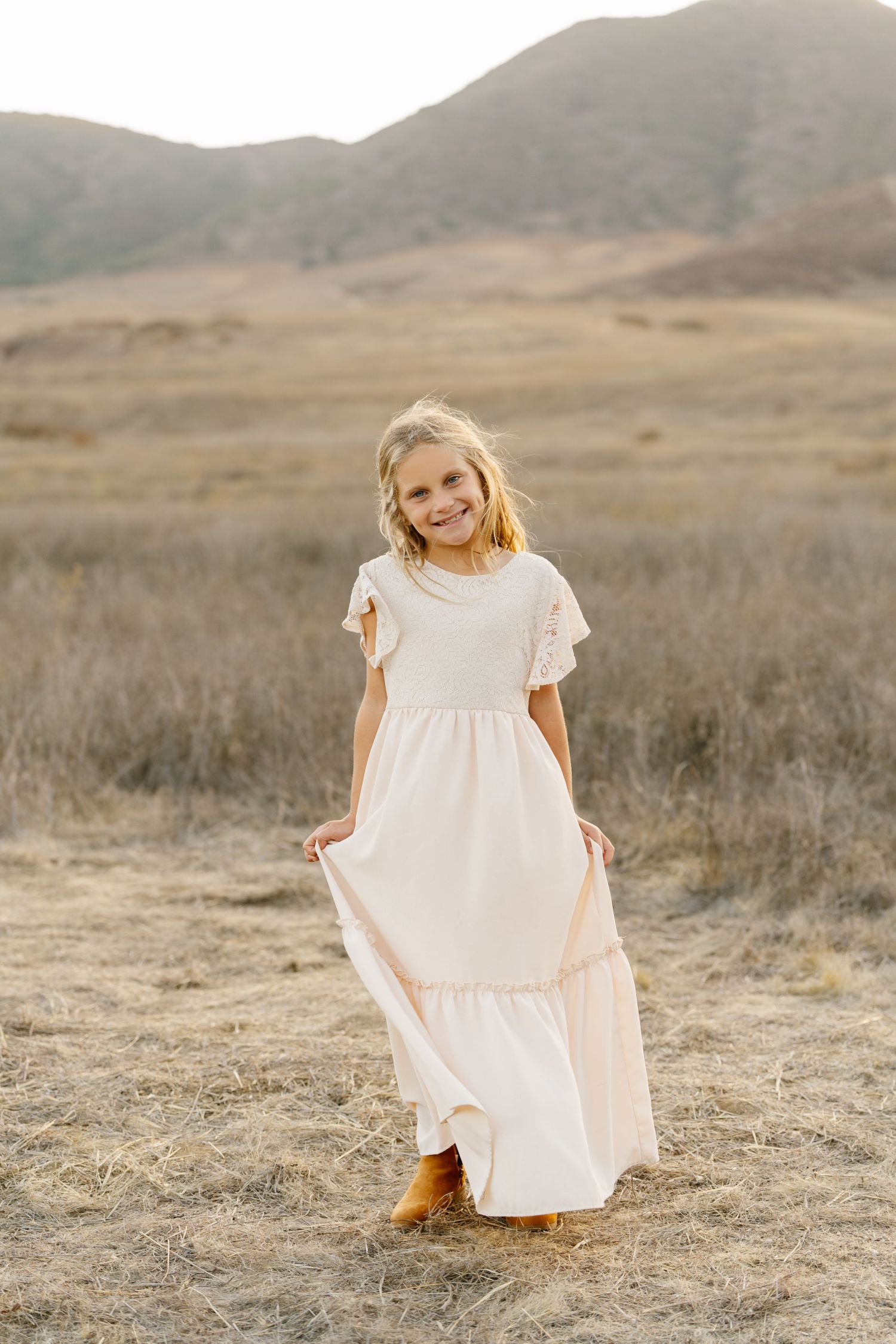A smiling young girl in a flowing cream maxi dress and cowboy boots stands in a golden field with mountains behind her.