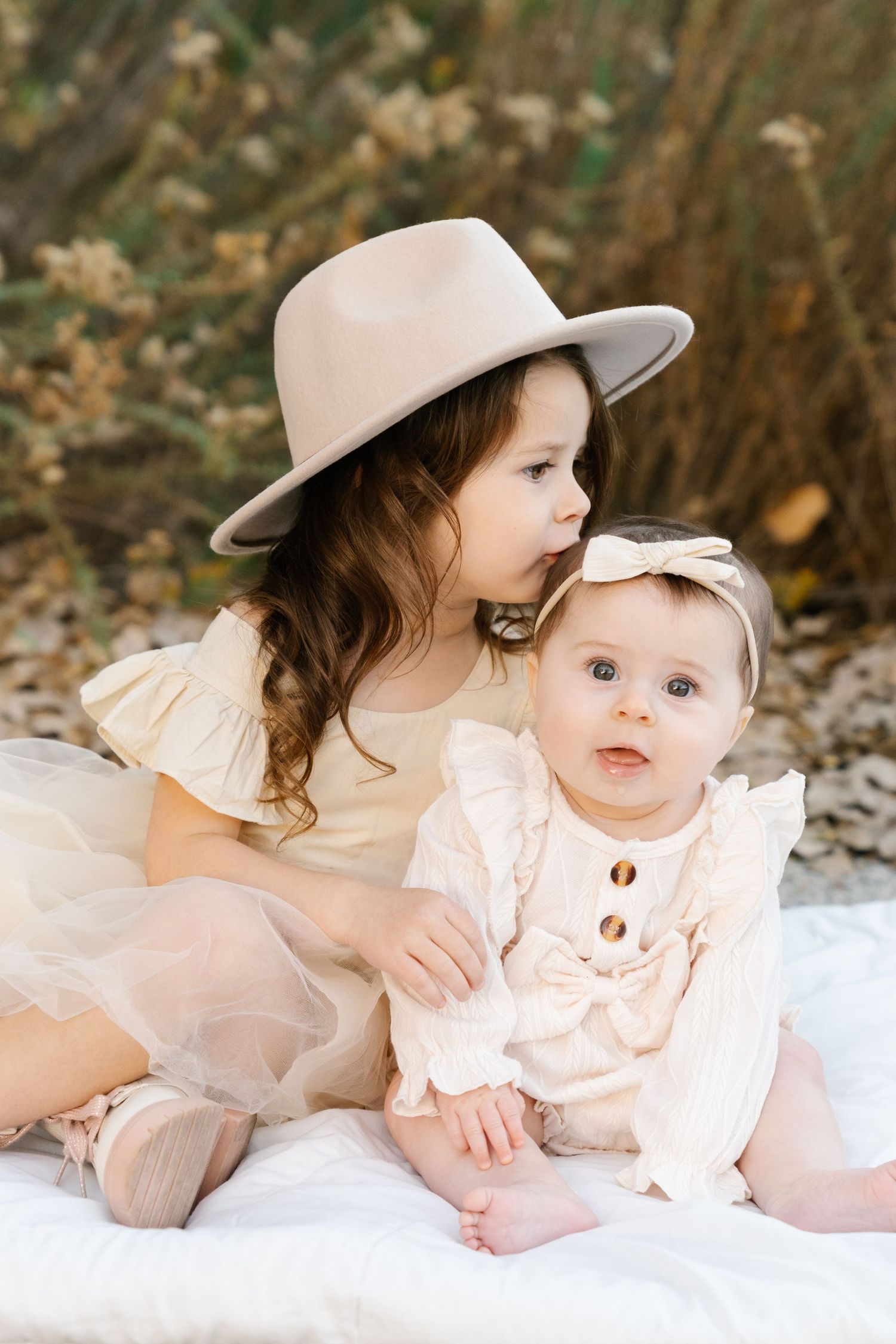 Two children in cream colored outfits sit together on white blanket outdoors.