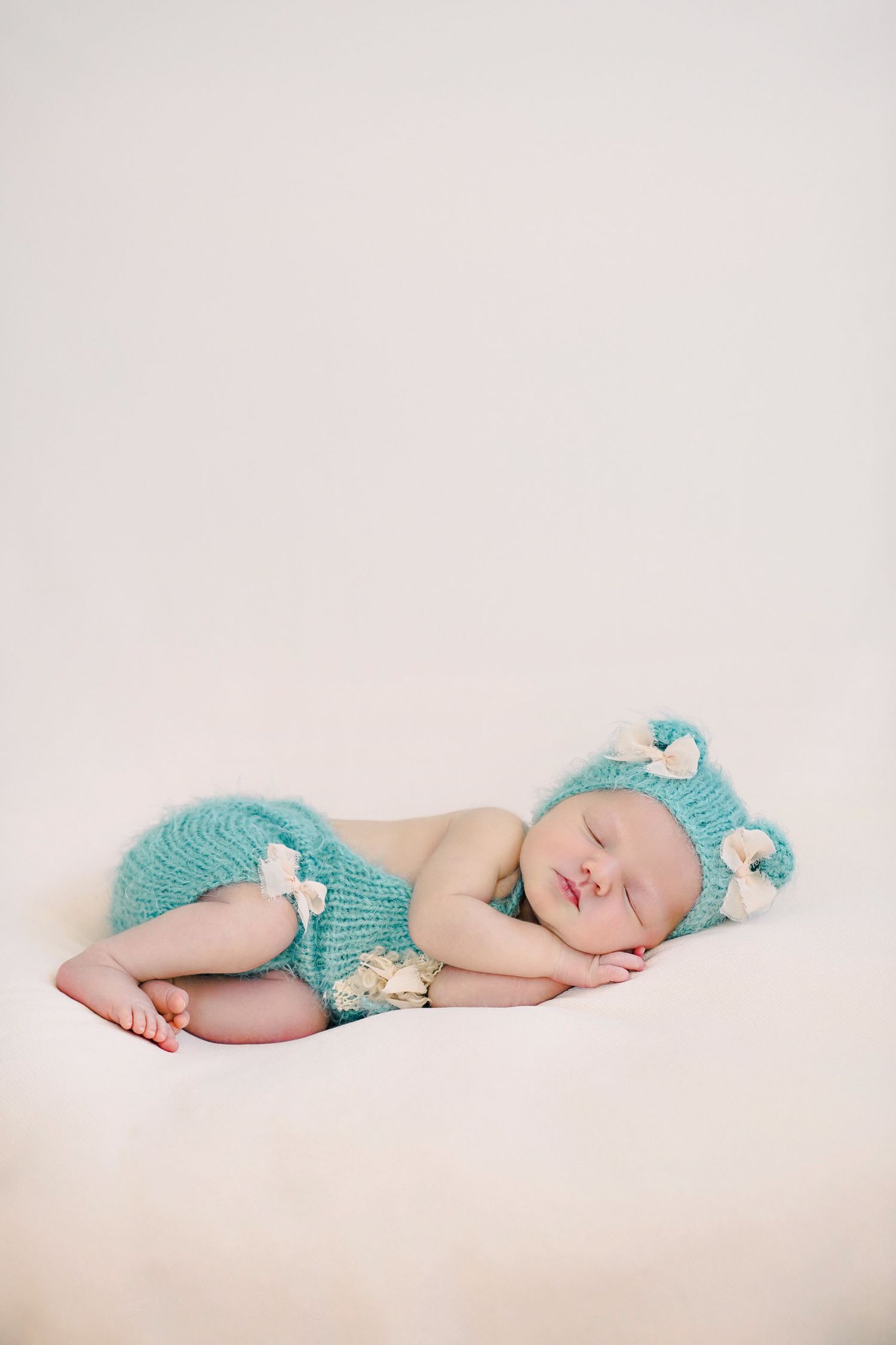 Sleeping newborn wearing turquoise knit bear outfit and matching hat on beige background.