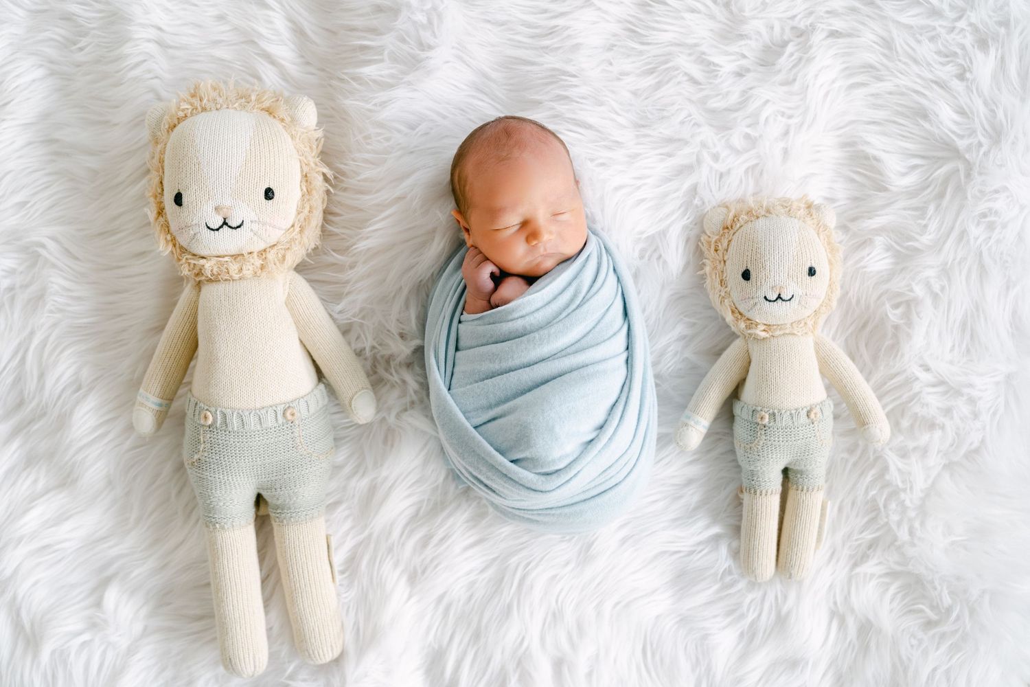 Swaddled sleeping baby between two plush lion toys on white furry rug.