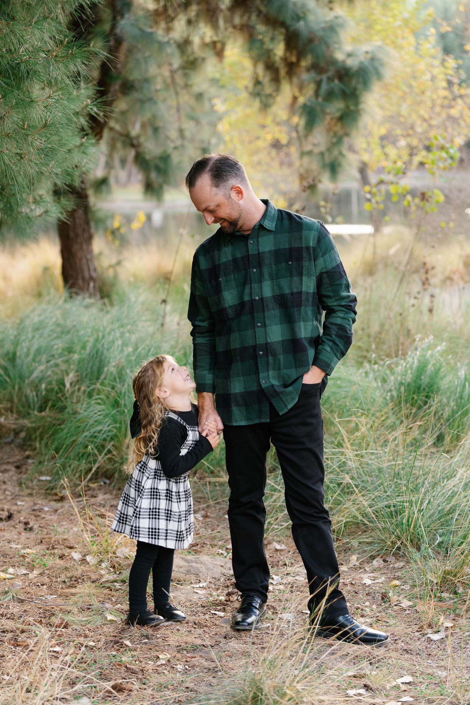 Parent and child share a sweet moment outdoors among tall grass and evergreen trees.