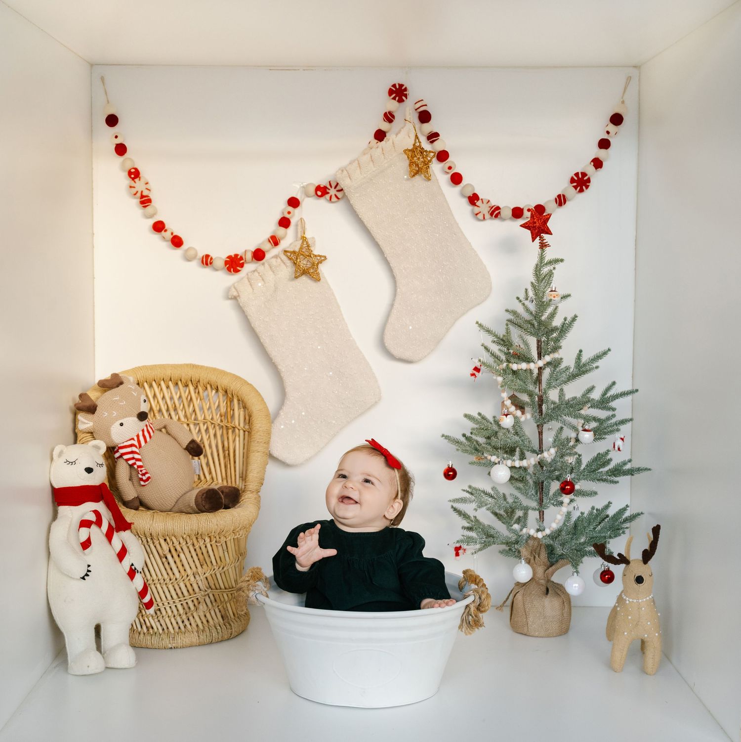 Baby in white tub with Christmas stockings, mini tree, stuffed animals, and red garland holiday backdrop.