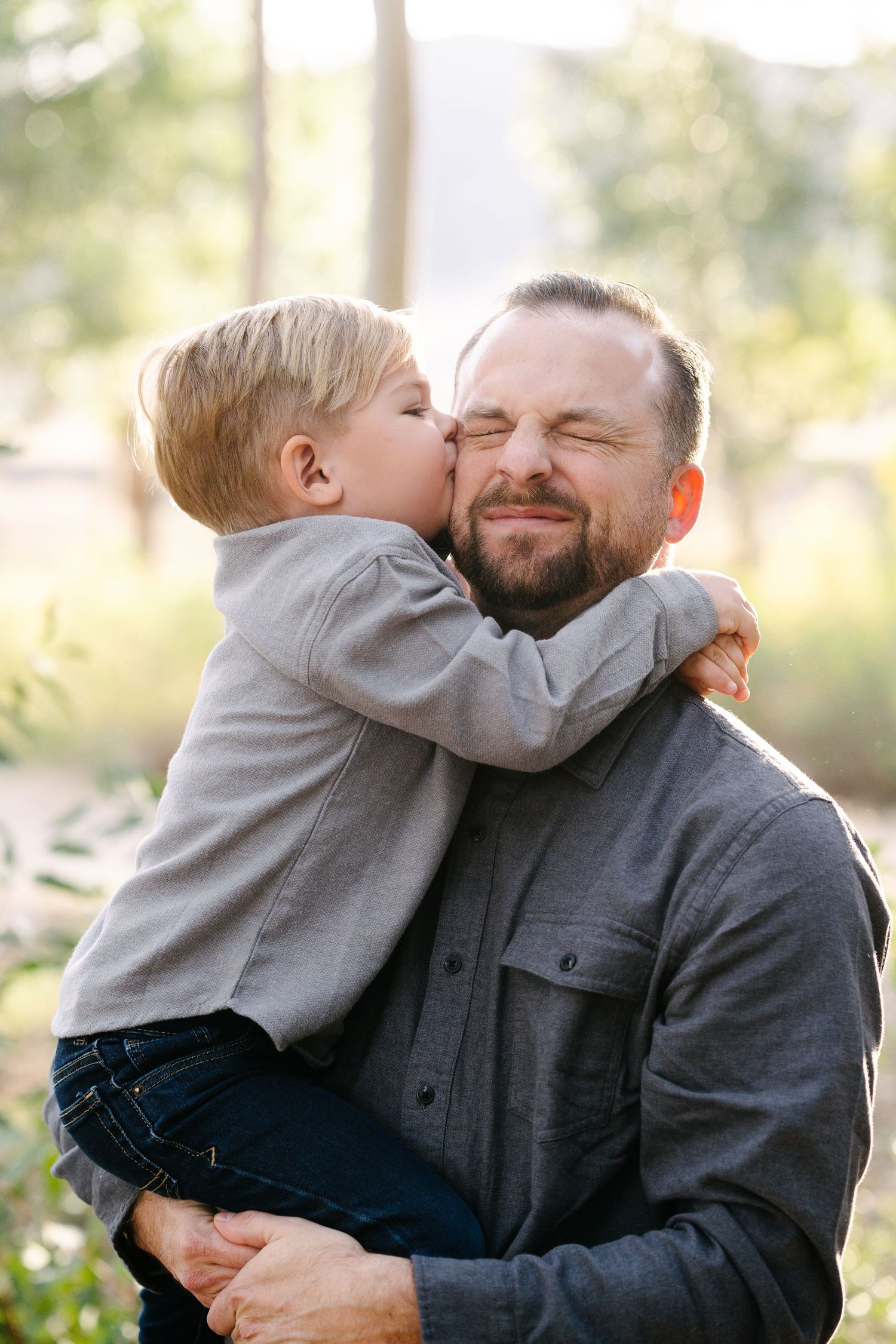 A young boy kisses his bearded father on the cheek while being held outdoors in a sunlit garden setting.