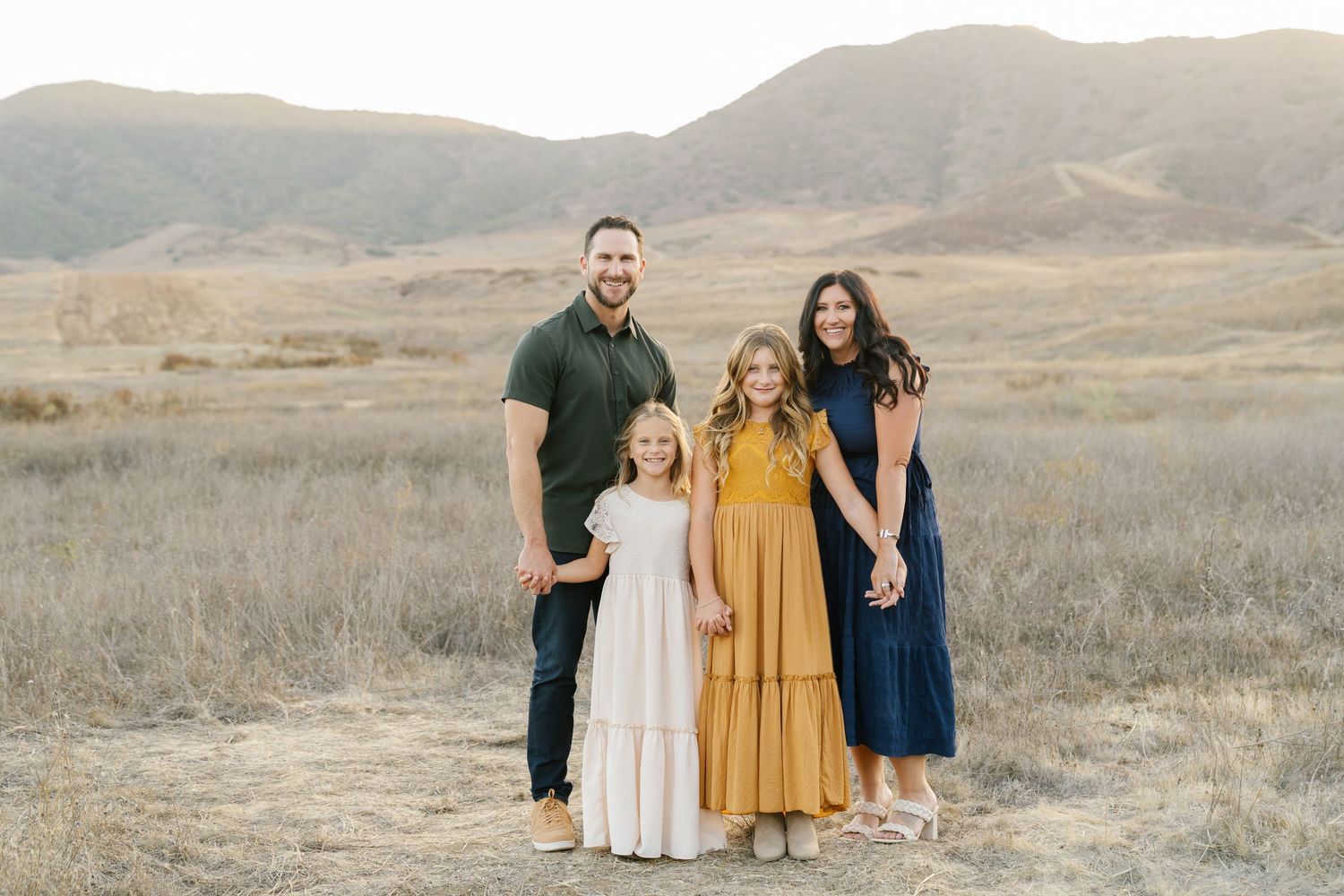 Family of four smiling in a golden field with mountain backdrop during sunset portrait session.
