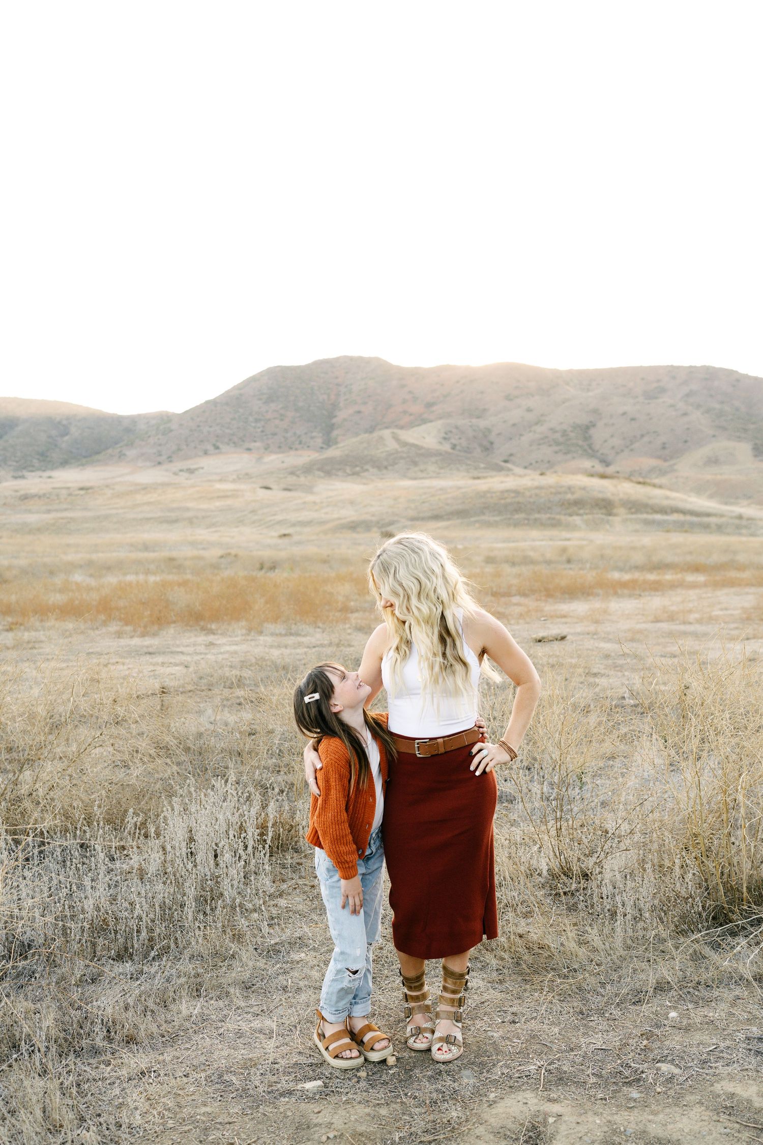 A young girl kisses a blonde woman in a red skirt standing in a golden field with rolling hills backdrop.