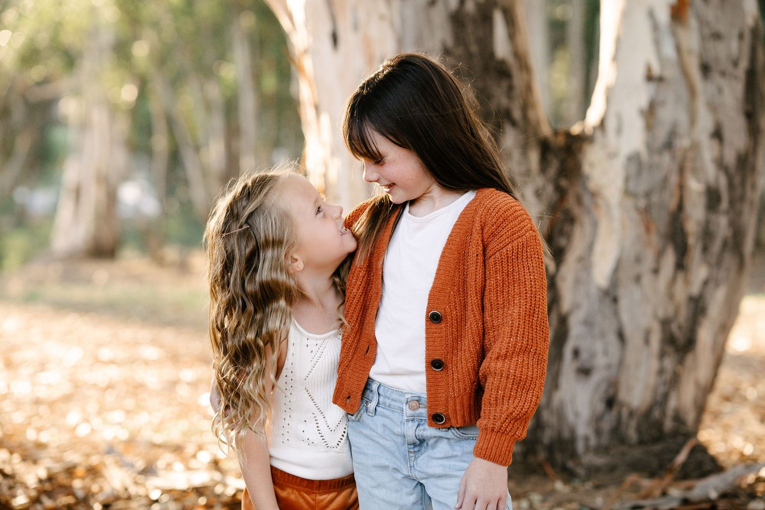 Two young girls in autumn outfits smile at each other among fall leaves and trees.