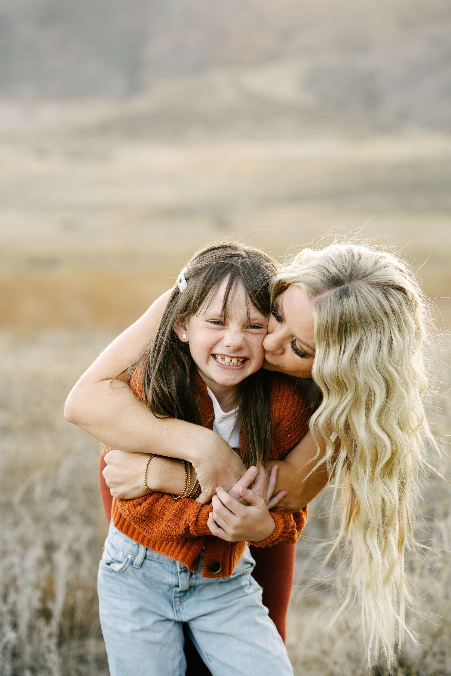 A blonde mother hugs and kisses her smiling young daughter outdoors in a golden field during autumn.