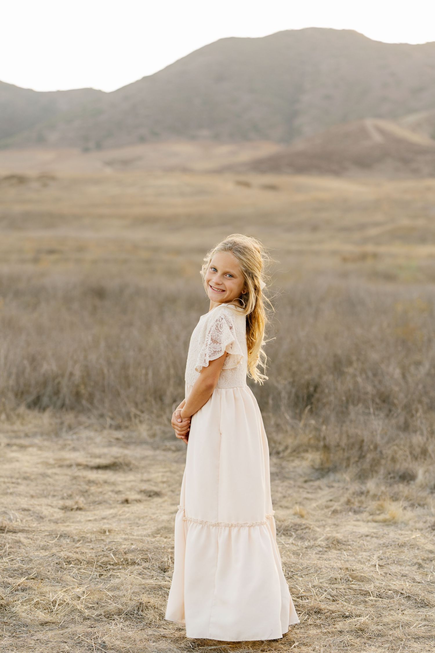 A young girl in a long white lace dress stands in a golden field with rolling hills behind her.