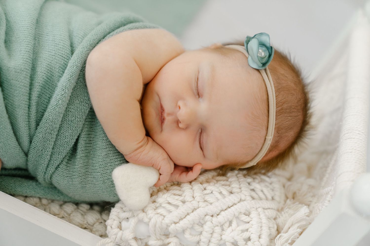 Peaceful sleeping infant in sage green wrap with teal headband on knit blanket.