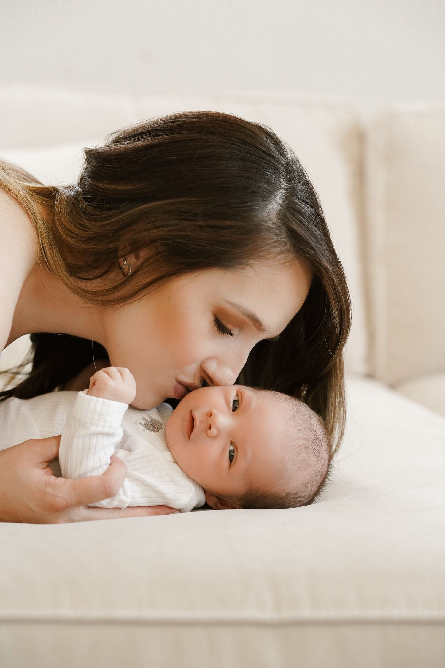 Tender moment between mother and baby lying on white couch.