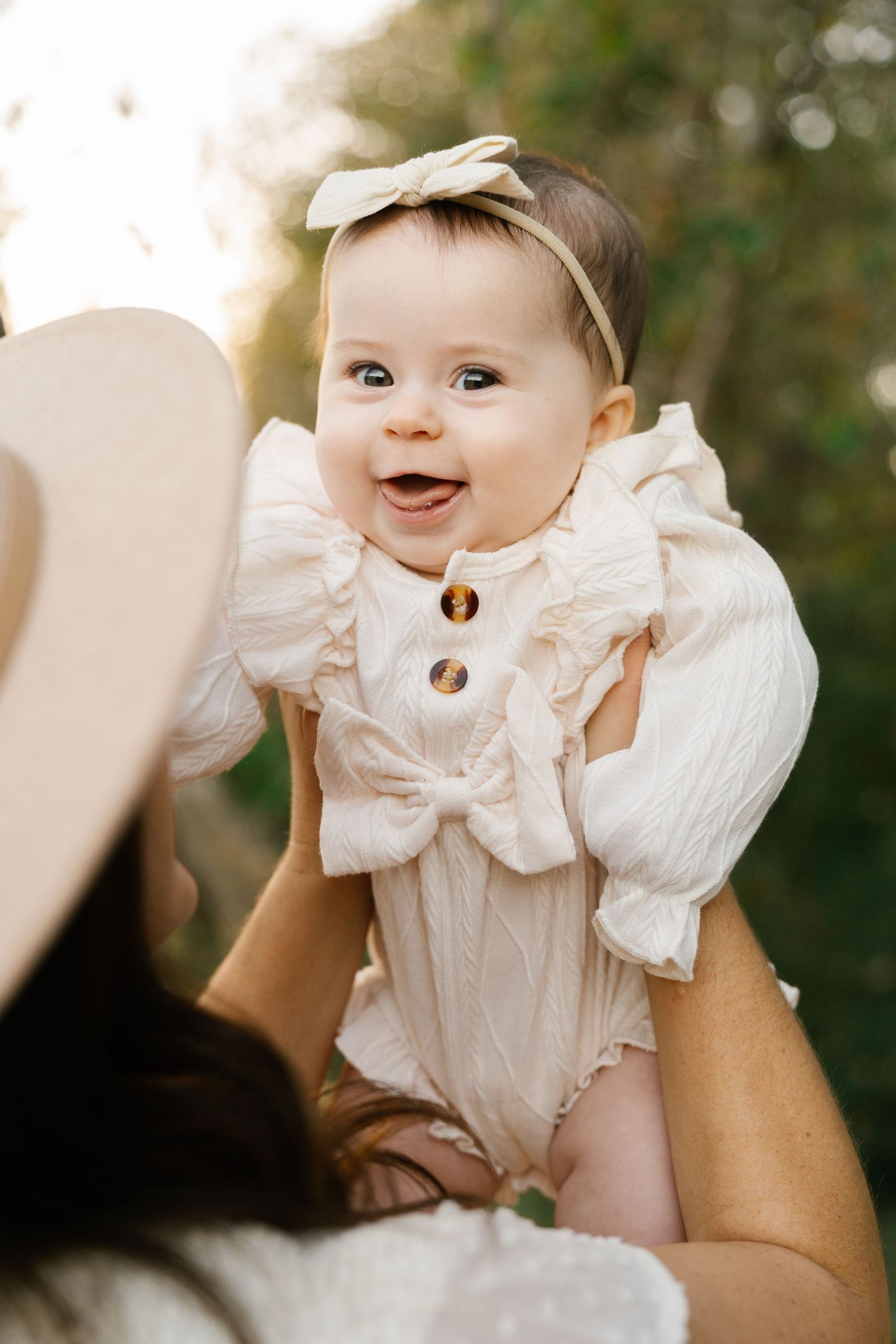 Baby wearing cream colored ruffled outfit with bow headband smiles brightly at camera.